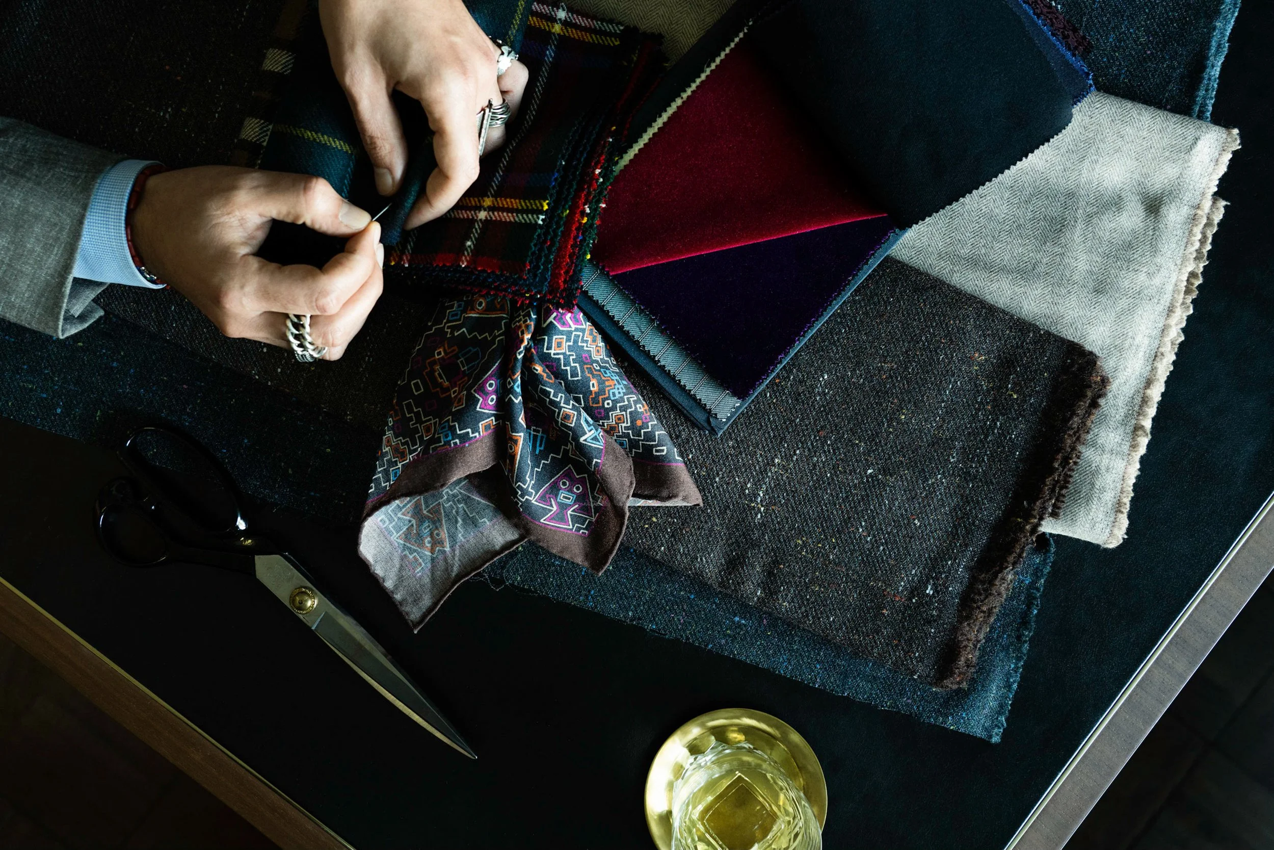 Person sewing fabric with plaid pattern on a table covered with various fabric swatches, with a glass of white wine nearby.