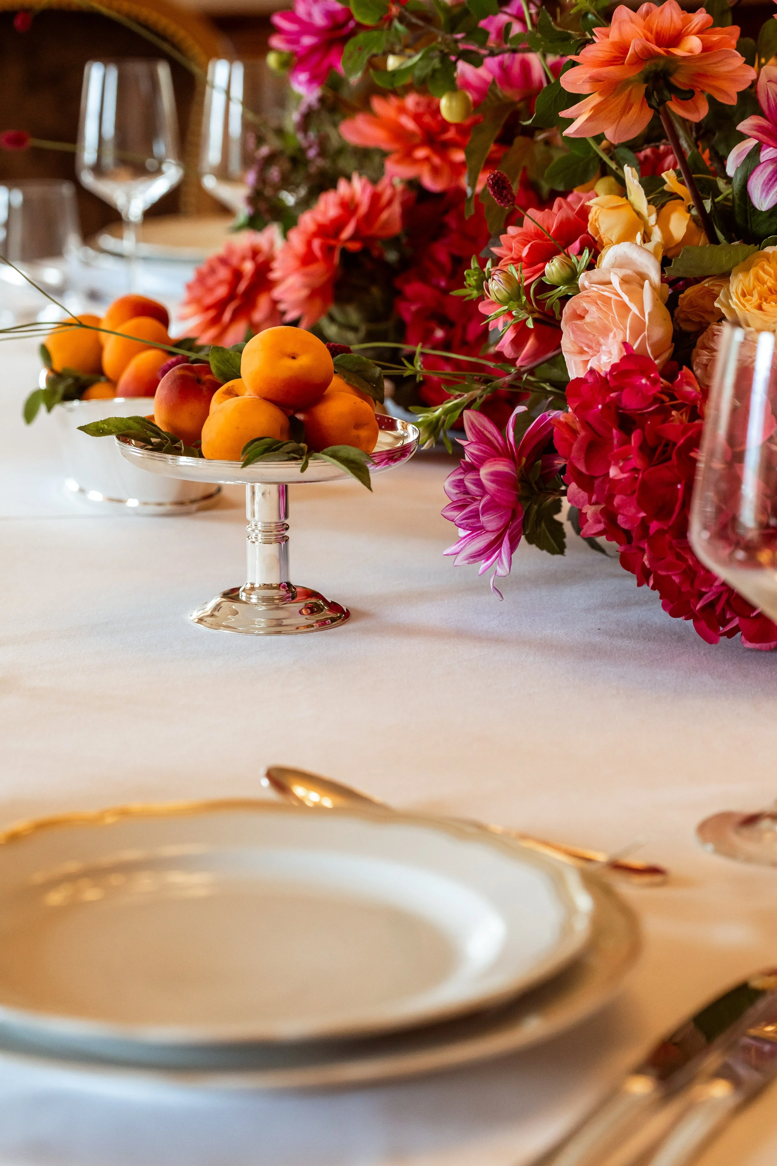 A table setting with a gold-rimmed white bowl in the foreground, pink and orange flowers in the background, a glass dish with peaches, and wine glasses on a white tablecloth.