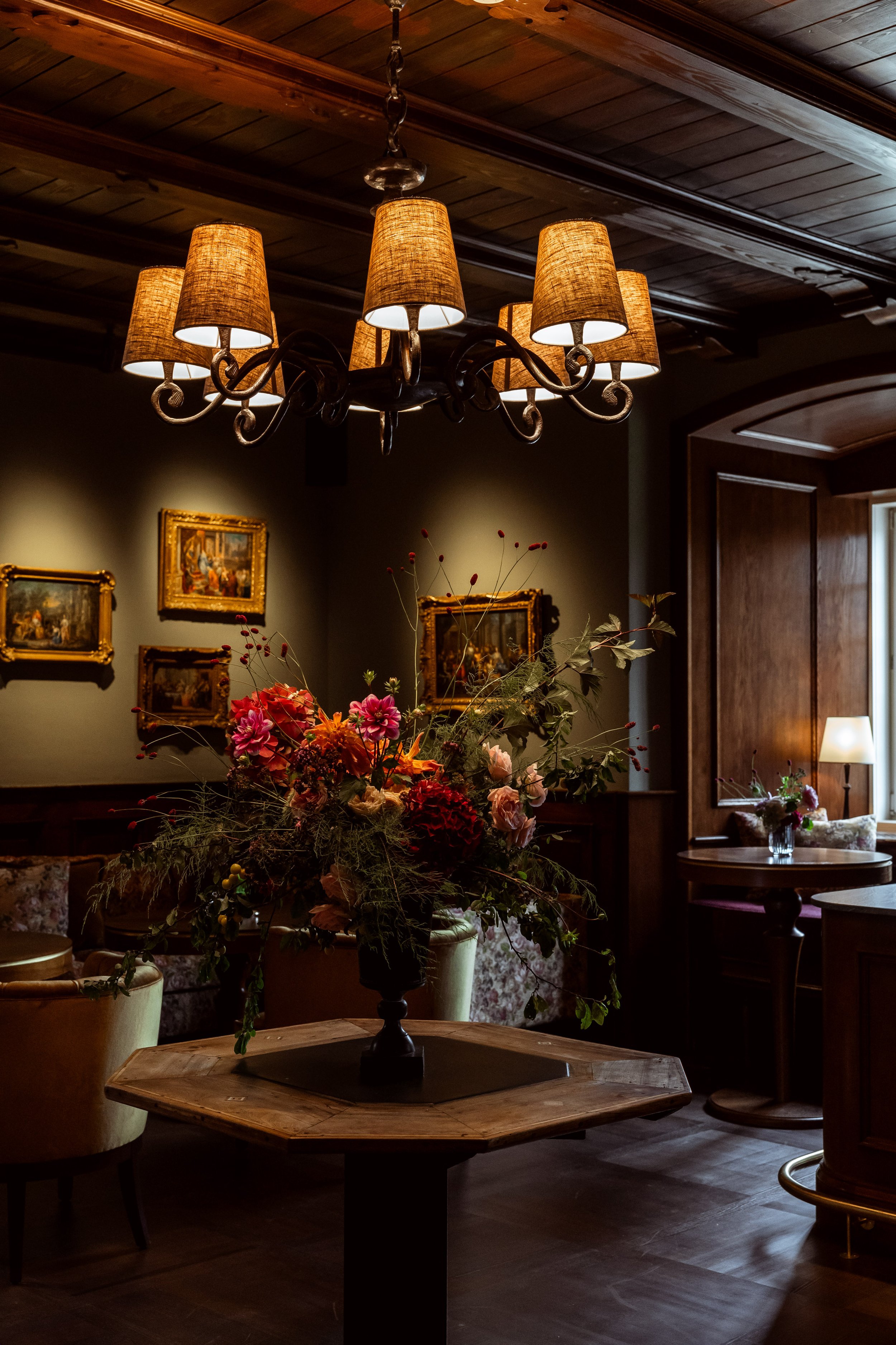 A chandelier with six warm-lit lampshades hanging from a dark ceiling in a cozy, elegantly decorated room with framed paintings and a flower arrangement on a table.