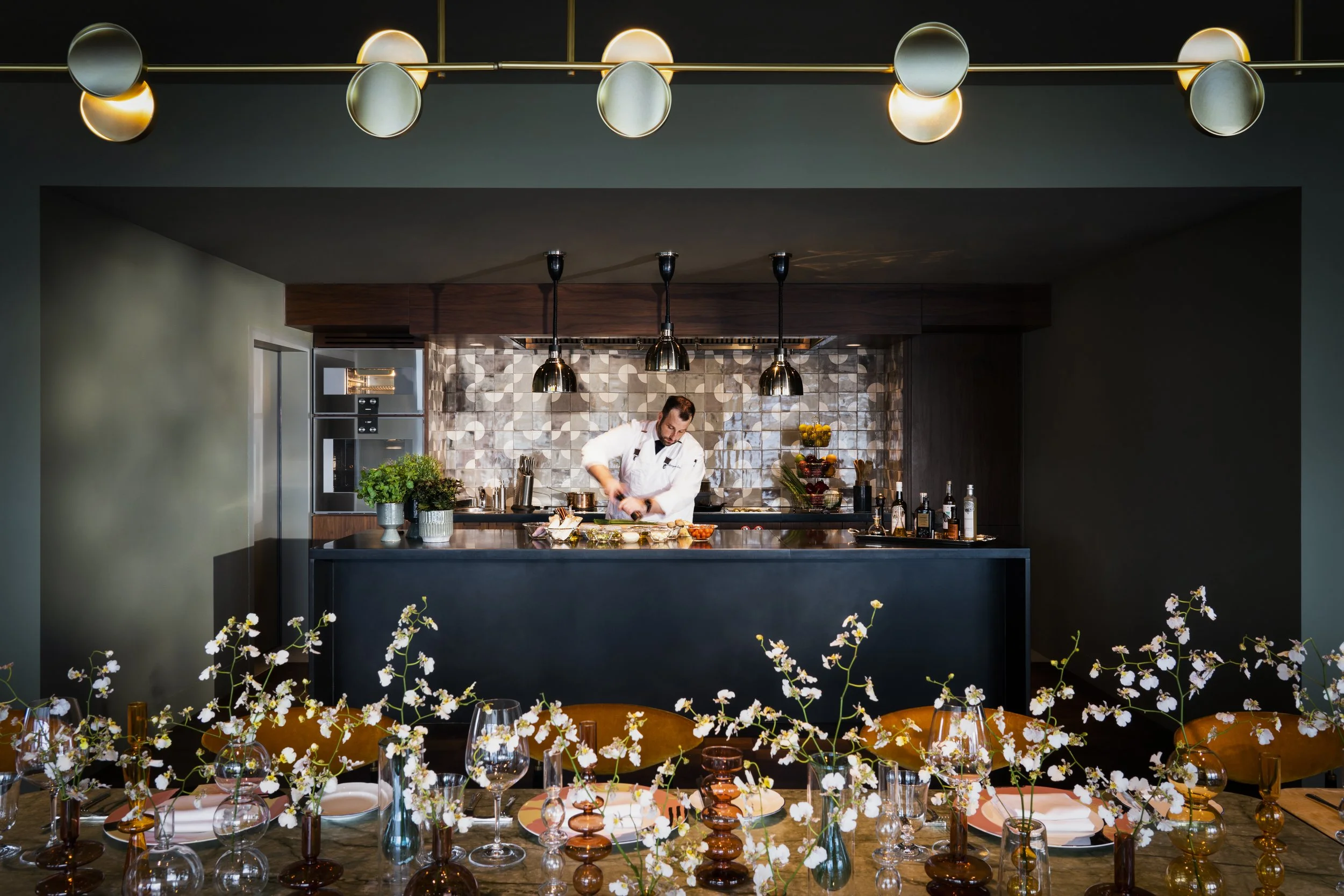 A chef prepares food in a modern kitchen with hanging pendant lights and a patterned backsplash, seen from a dining area with floral centerpieces and glassware on a table.