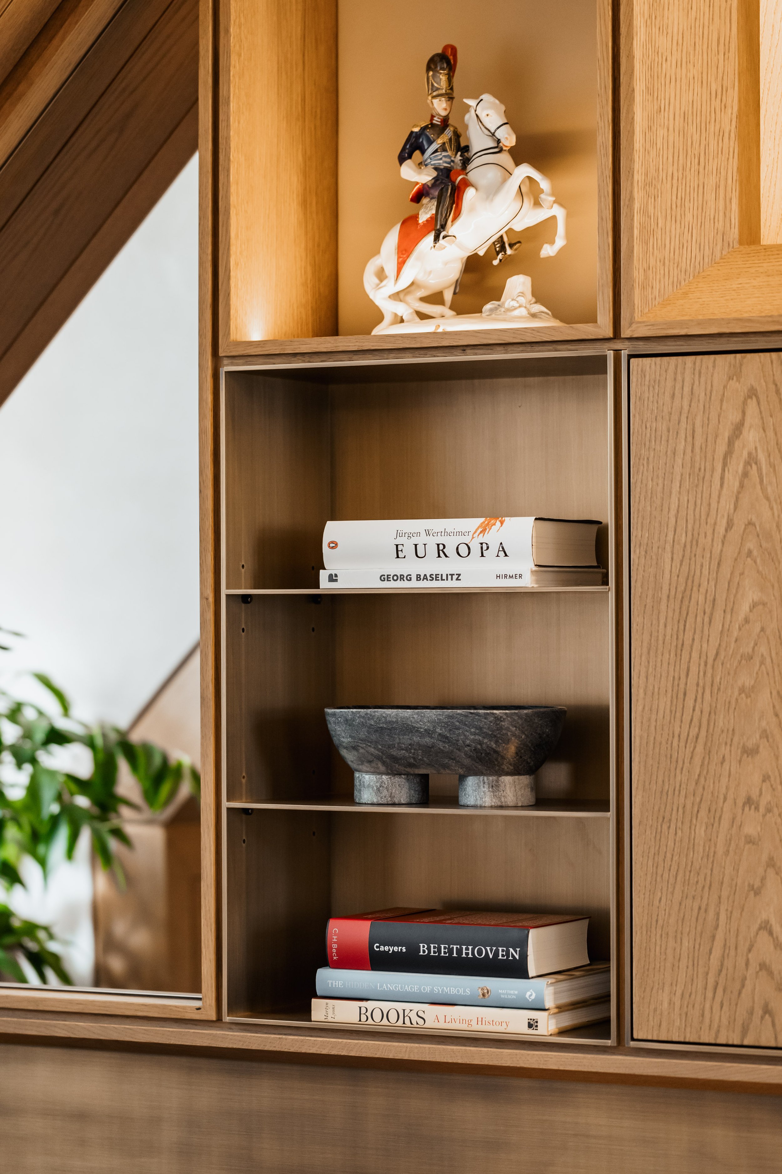 A wooden bookshelf with decorative items, including a porcelain figurine of a man on a horse, a few books, and a stone bowl.