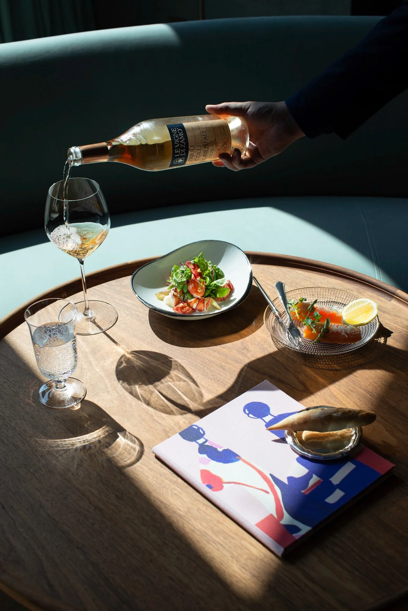 A wooden table set with a glass of white wine, a plate of salad, a small bowl of salmon with lemon, two empty glasses, a colorful napkin, and a menu with an abstract blue and pink design. A person is pouring wine from a bottle.