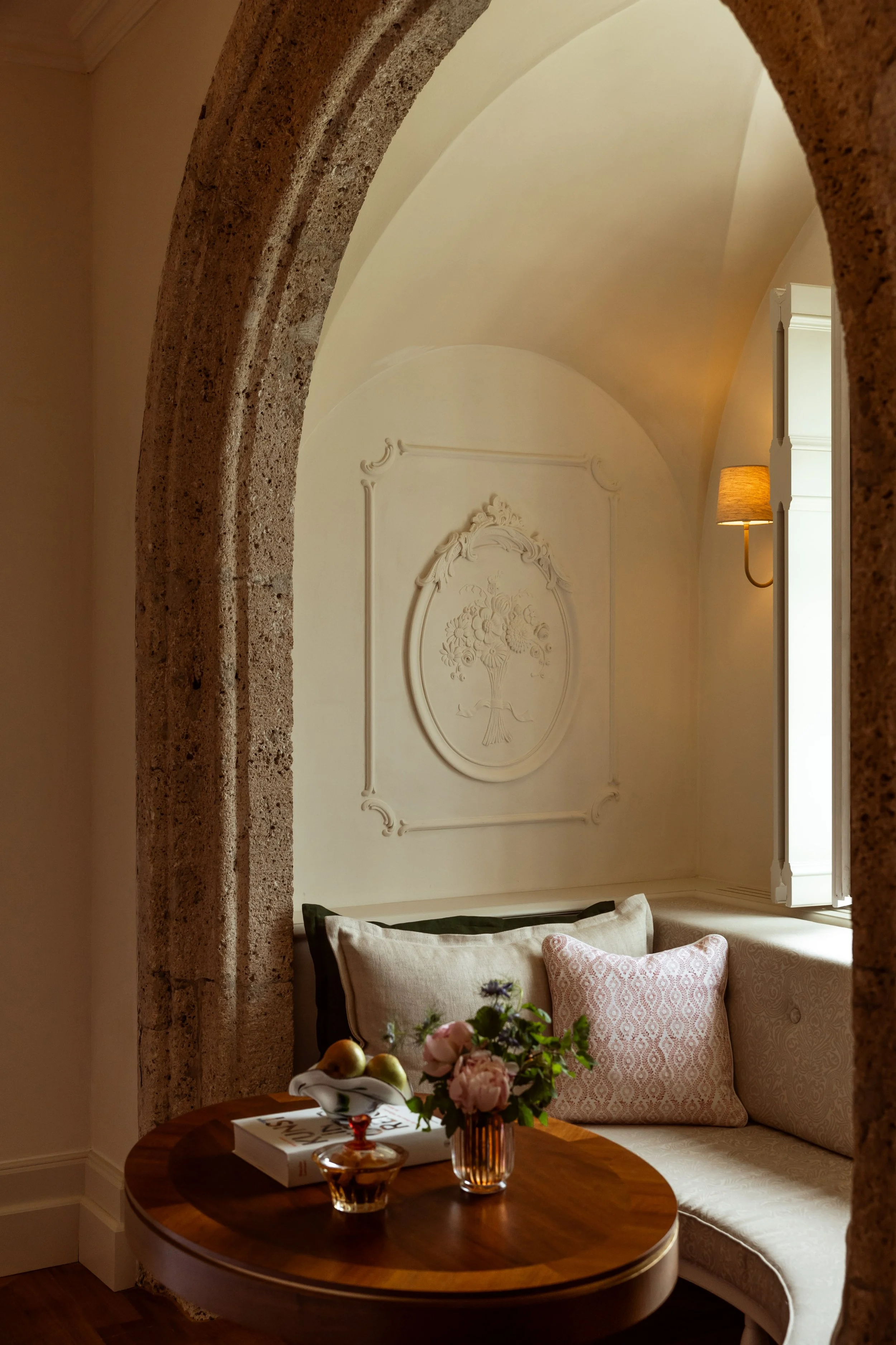 A cozy corner of a room with beige walls and a decorative white wall panel featuring a floral motif. A window with white shutters partially opened lets in natural light. There is a wall-mounted lamp with a brown lampshade. A curved upholstered bench 