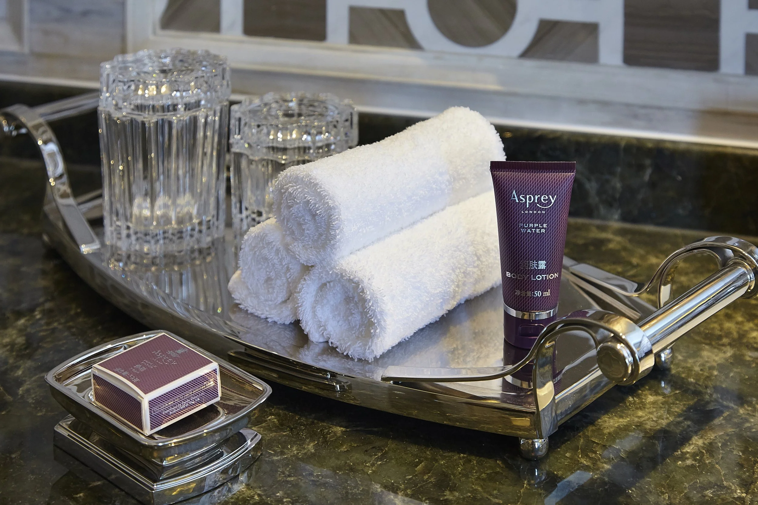 A silver tray on a dark green marble countertop holding a stack of three clear glass drinking glasses, a folded white towel, a purple bottle of lotion labeled 'Asprey London Purple Water', a small deck of playing cards, and a pair of silver tongs. In
