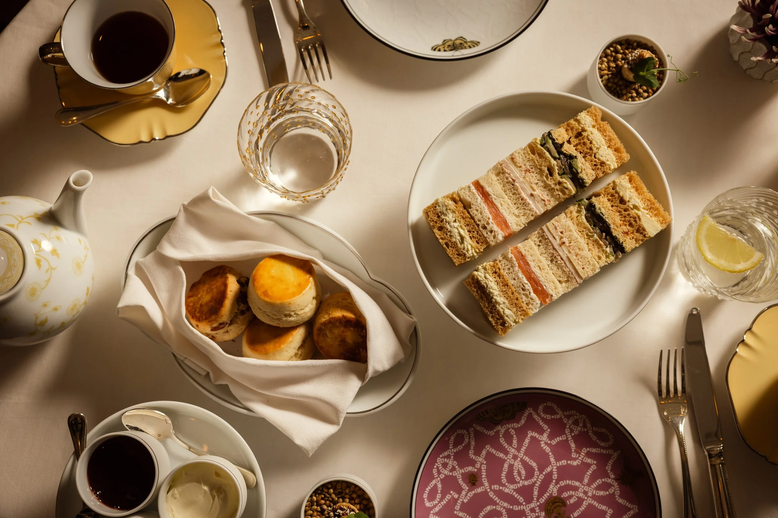 A table set with tea, water, and coffee, a plate of assorted finger sandwiches, a basket of small bread rolls, a teapot, a cup of tea, and small bowls of cream and jam, decorated with flowers and napkins.