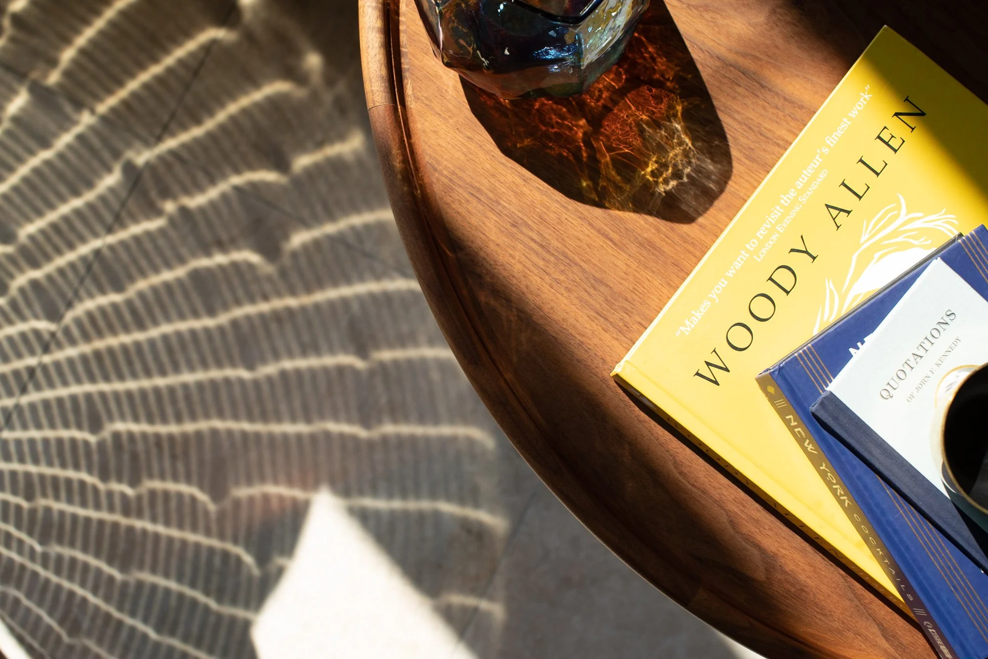 Close-up of a wooden table with two books and a glass of dark beverage casting a shadow on the table. Part of a person's striped sleeve is visible on the left side.