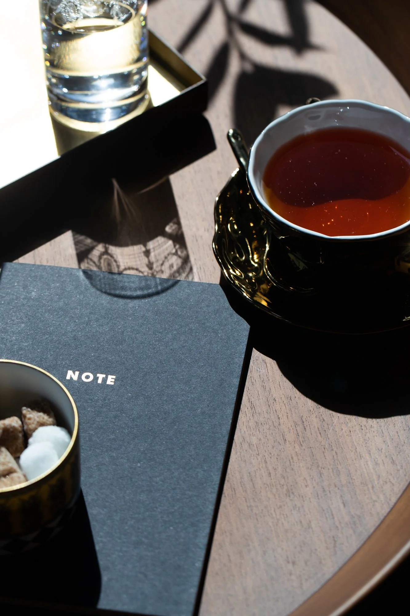 A top-down view of a table with a glass of water, a cup of tea in a black and gold ornate cup, a dish with cookies, and a notebook with the word "NOTE" on the cover.
