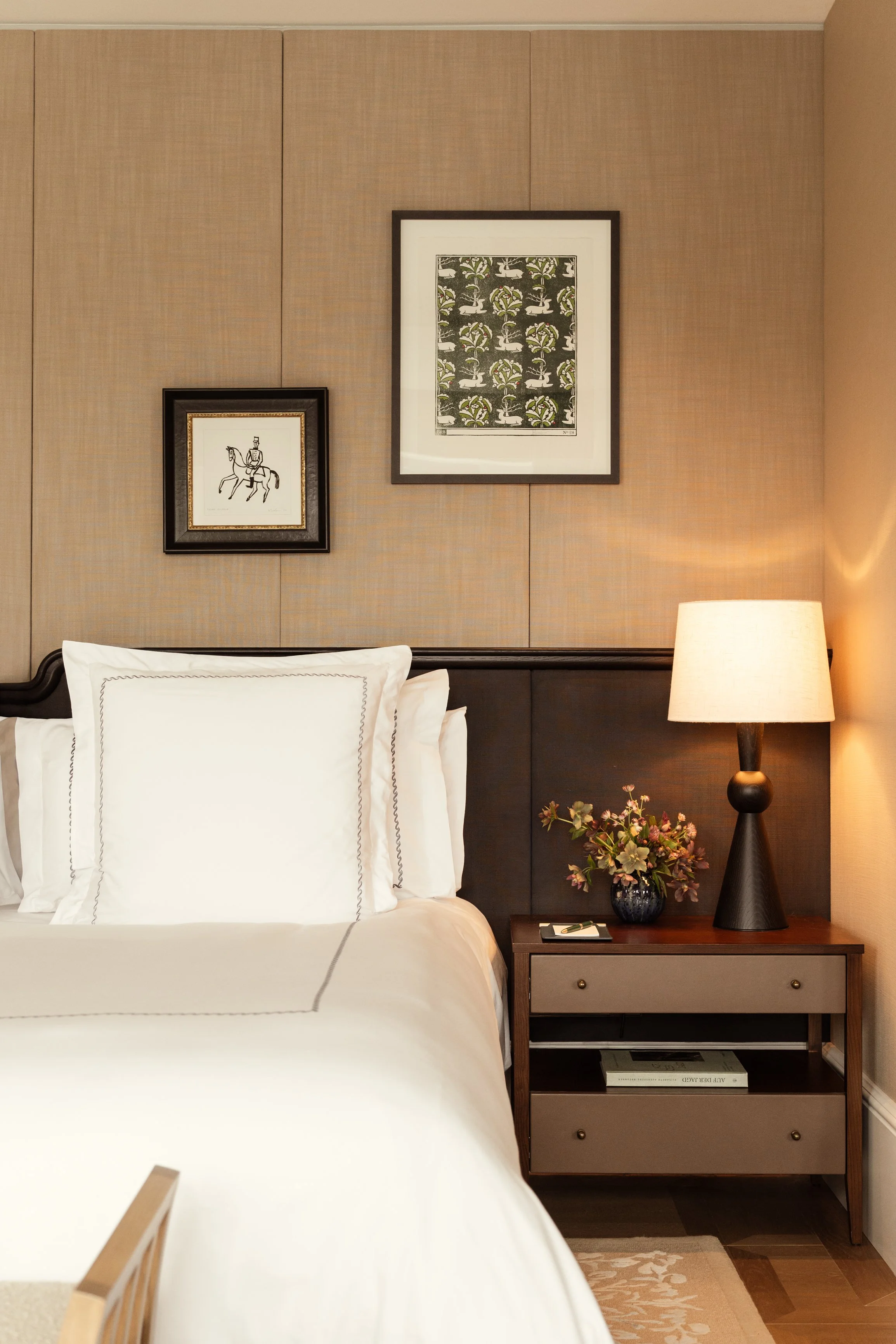 A bedroom with a bed, two white pillows with black stitched borders, two framed pictures on the beige wall behind the bed, a beige nightstand with a black table lamp, a vase with flowers, and a drawer with a book titled 'AU DÉPART'.