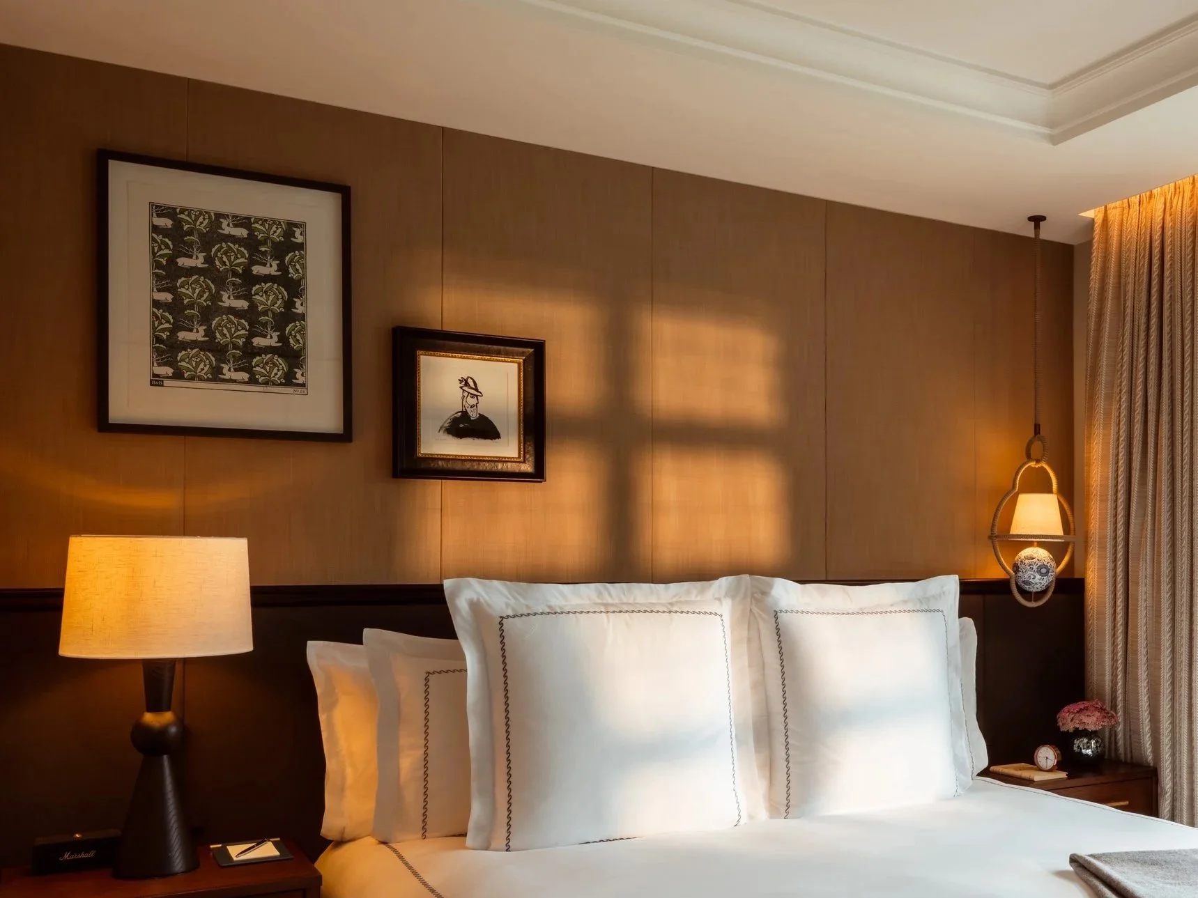 A cozy bedroom with a wooden paneled wall, white bed with decorative pillows, a bedside table with a lamp, framed artwork on the wall, and a decorative hanging lamp next to curtains.