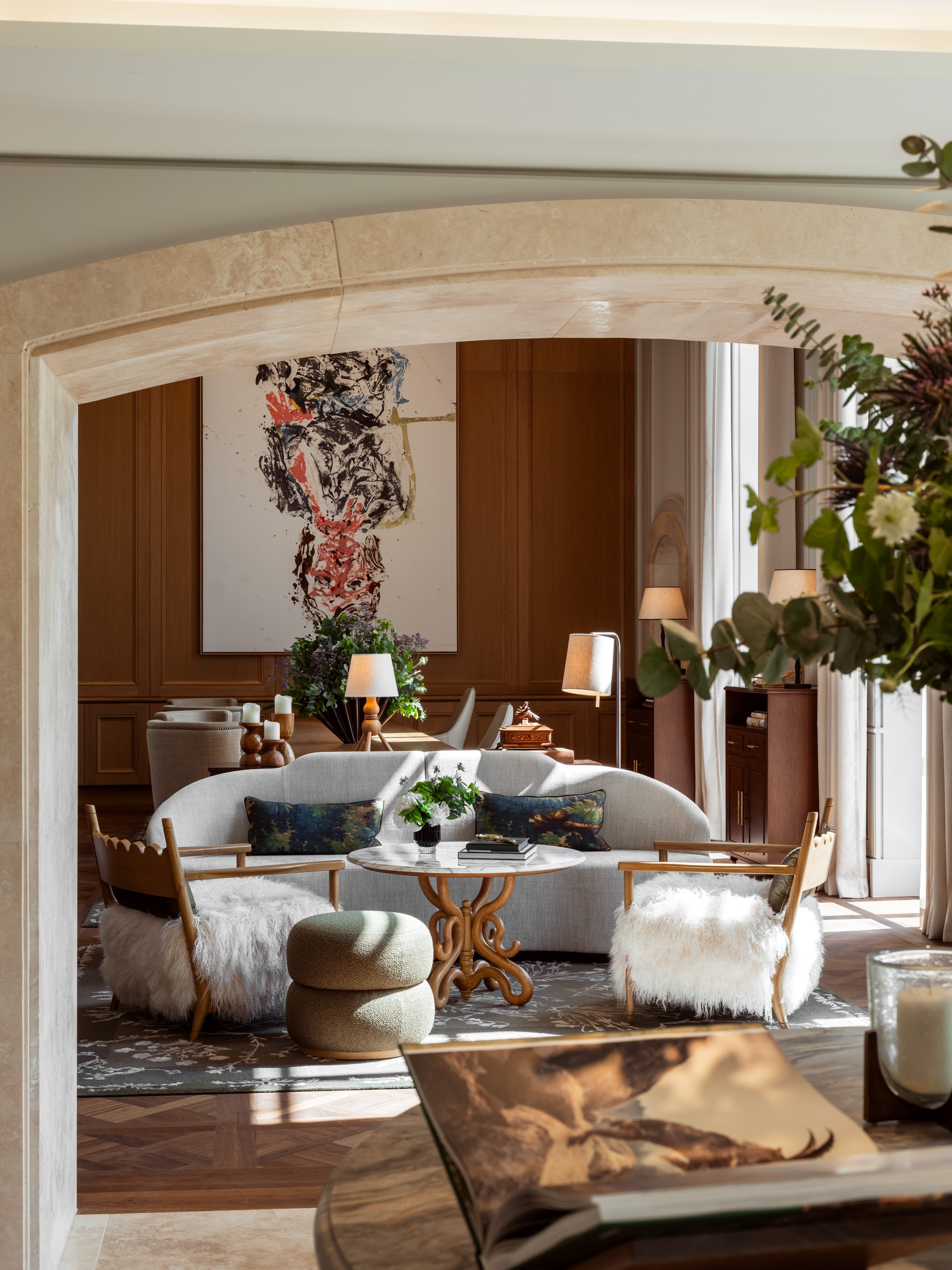 A cozy living room with a cream-colored sofa, fluffy white ottomans, wooden chairs with cushions, a round marble table with flowers, lamps, and artwork on wood-paneled walls, illuminated by natural light from large windows.