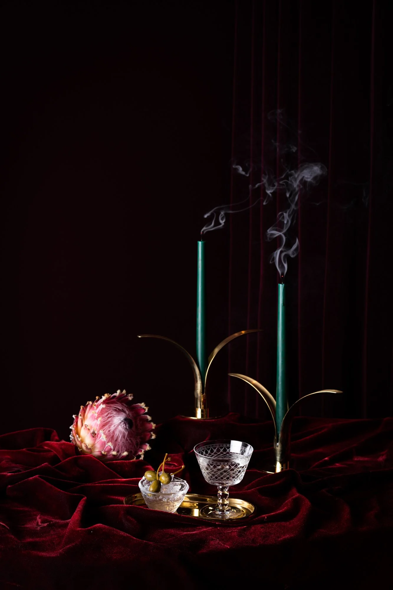 A still life scene with a dark background, featuring a flowering cactus, a glass bowl with white cherries, a designer crystal glass, a floral arrangement, and a bouquet of flowers, all placed on a red velvet fabric and illuminated by candle-like obje
