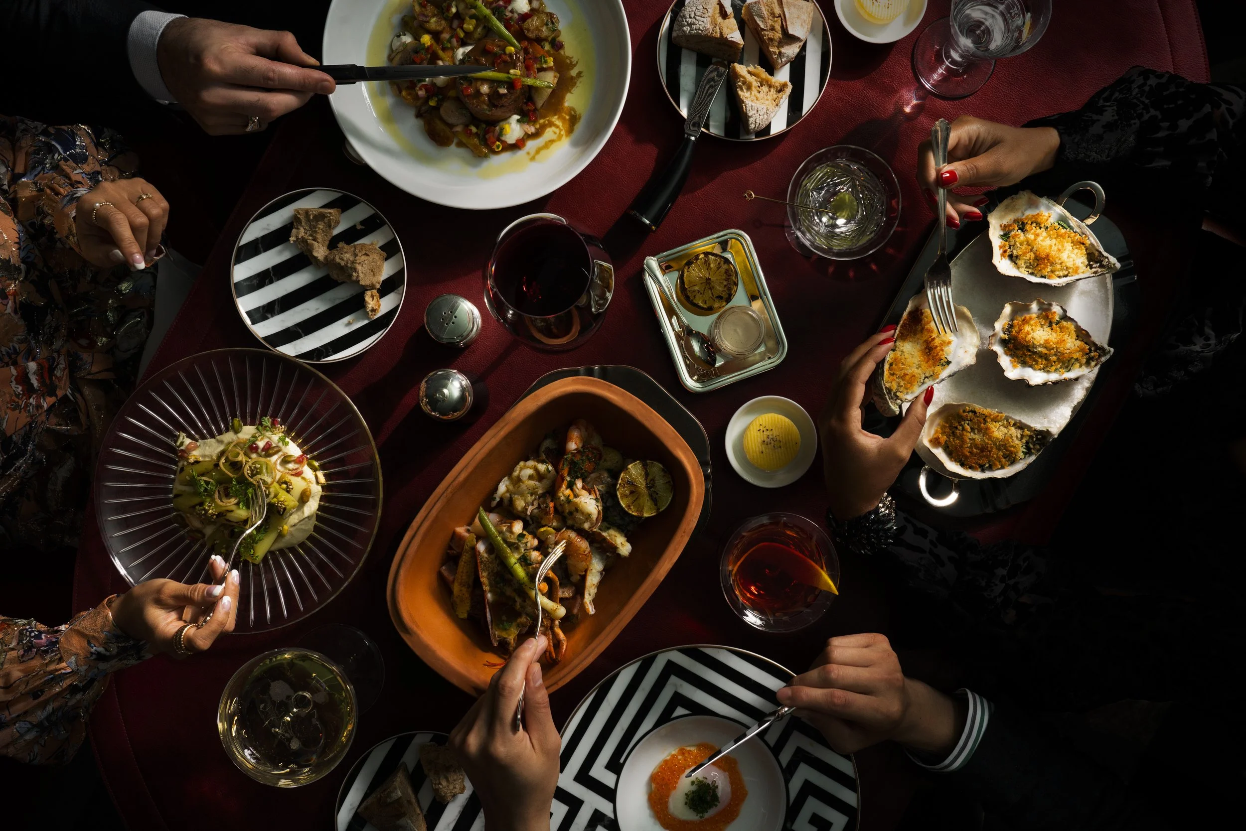 Top-down view of a dining table with various dishes including a bowl with salad, a plate with an egg dish topped with orange roe, a bowl of pasta, a platter of crostinis, and a bowl of chopped vegetables. People are reaching for the food with forks a