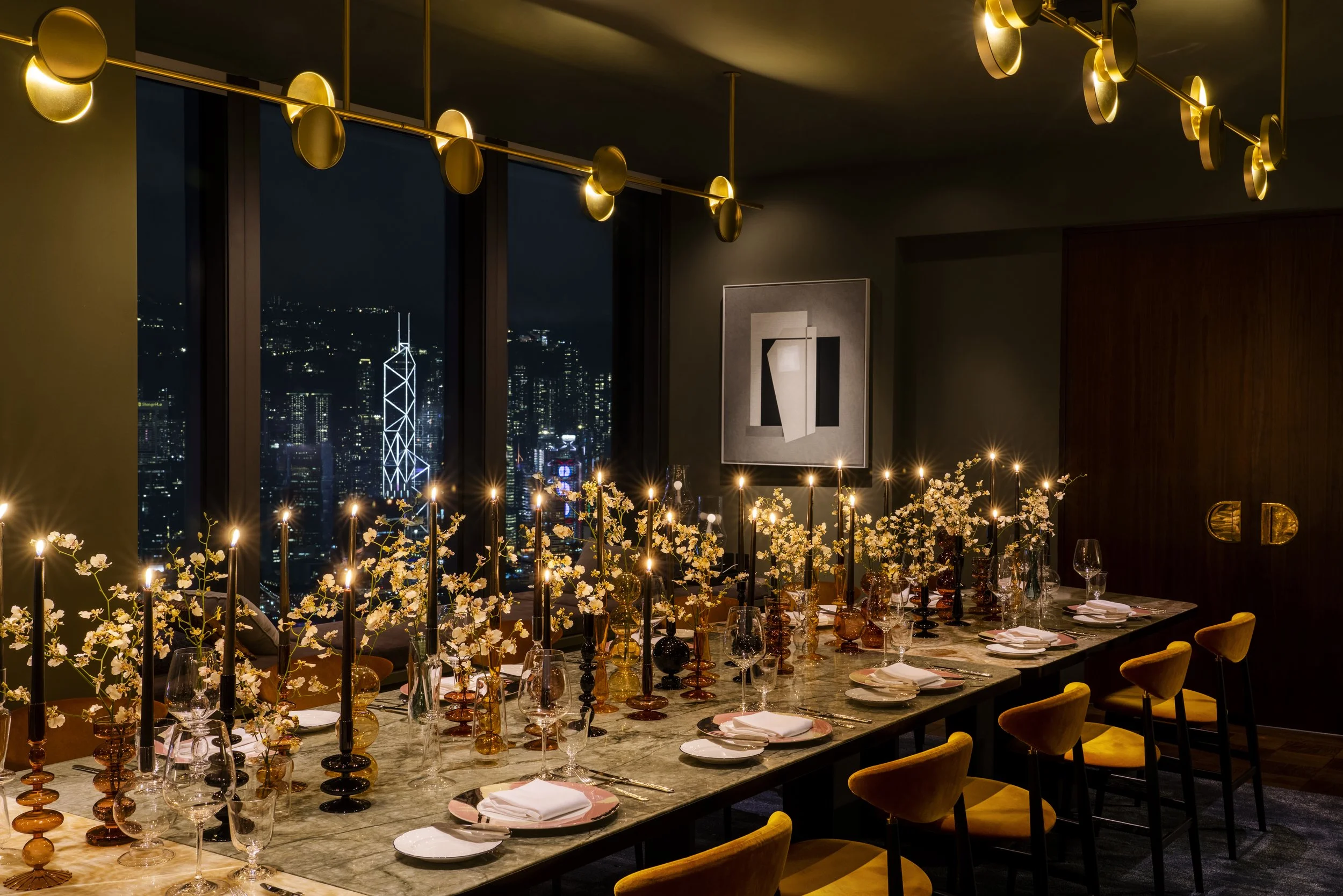 A luxurious dinner party setup with a long marble table decorated with flowers, candles, and glassware, overlooking a city skyline at night through large windows.