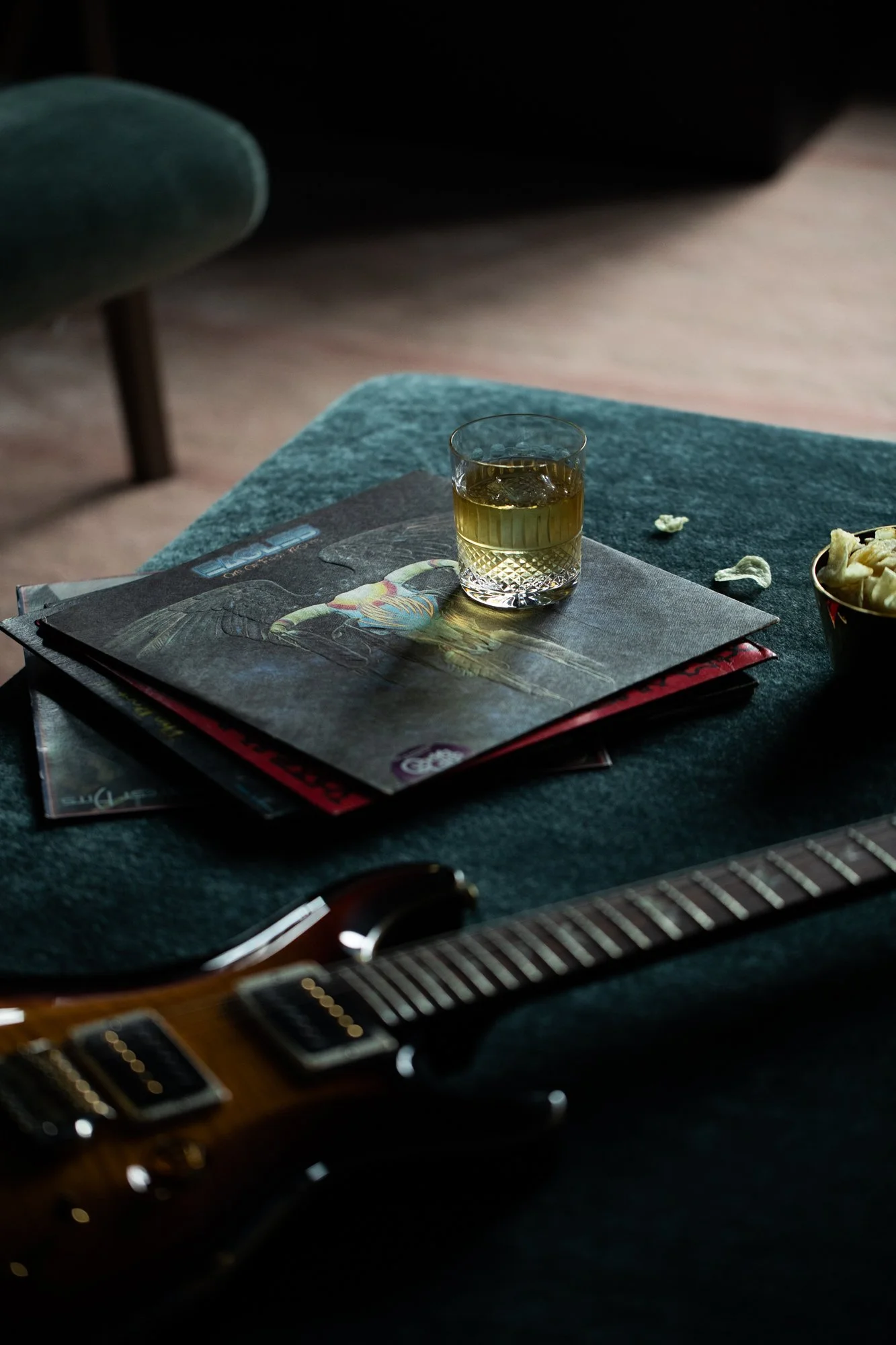 A glass of whiskey, a guitar, and magazines on a teal upholstered surface, with scattered popcorn and a bowl of popcorn nearby.