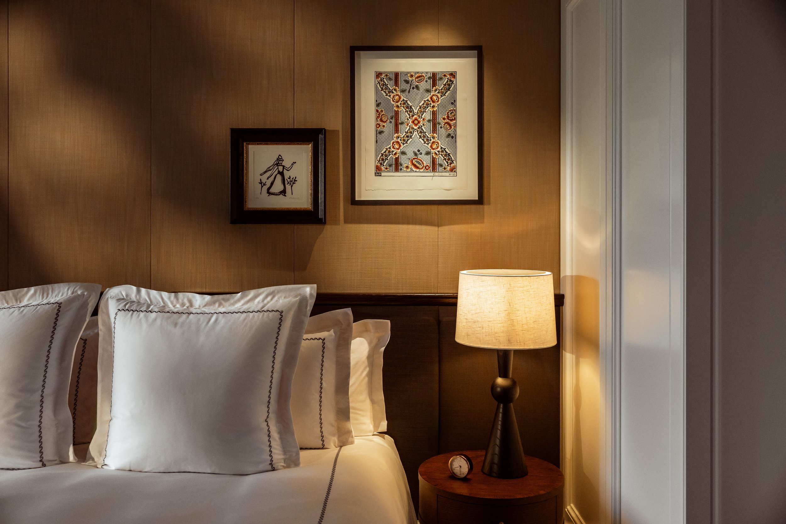 A bedroom corner with a neatly made bed featuring white pillows with dark piping, a wooden bedside table, a modern black lamp with a beige lampshade, a small clock, two framed artworks on a wooden-paneled wall, and a white-paneled door on the right.
