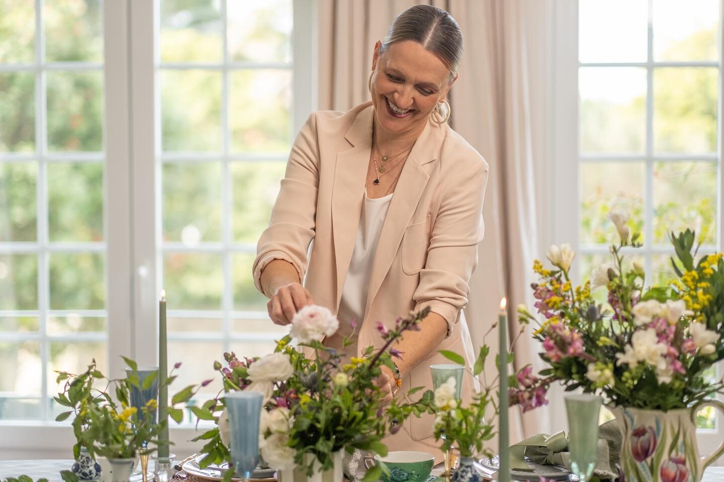 A woman arranging flowers on a dining table with large windows in the background.