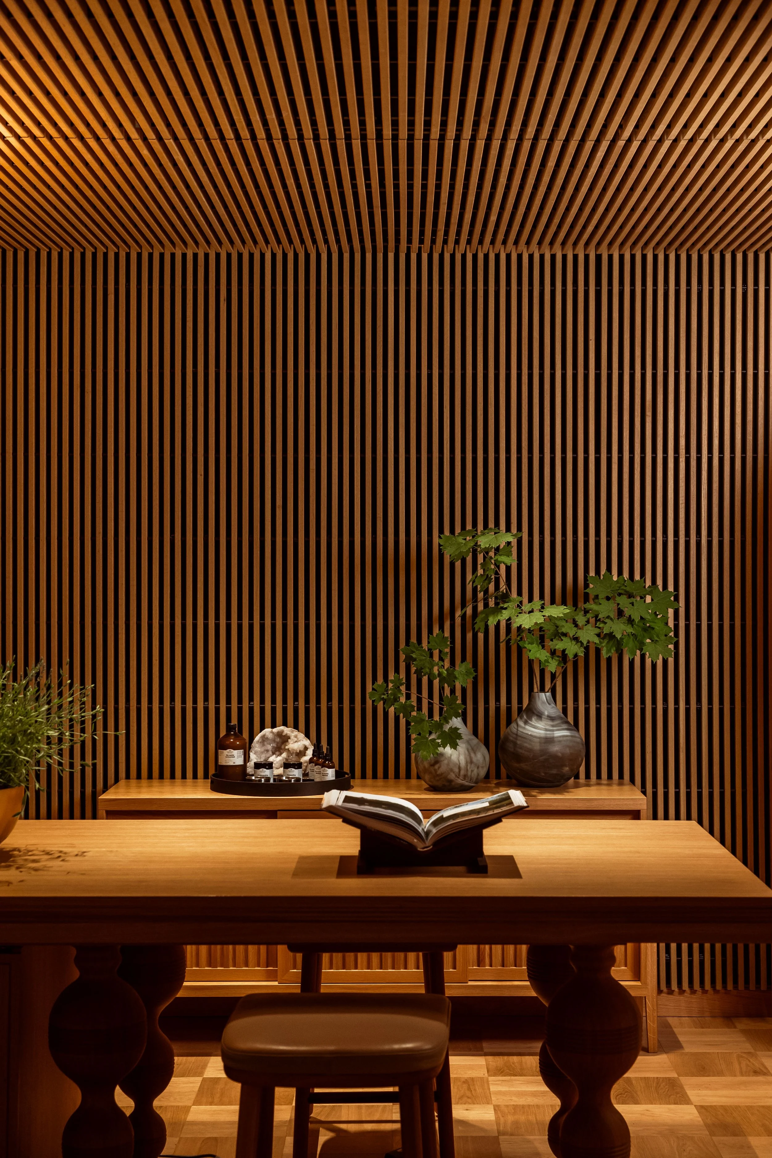 Interior scene with a wooden table, a small stool in front, decorative vases with green leaves, a tray with bottles and crystals, and a background of wooden slat wall.