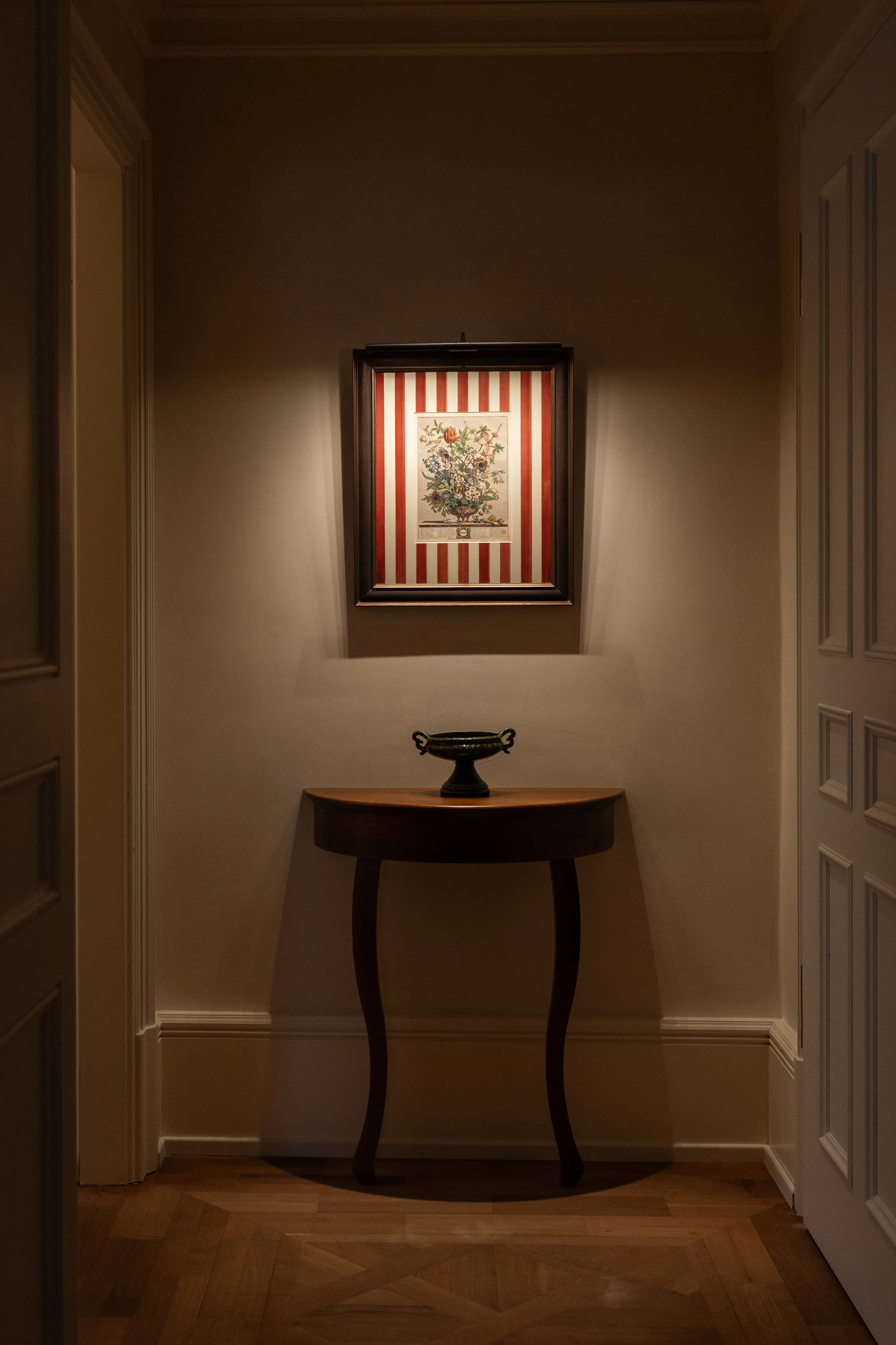 A framed botanical print hanging on a light-colored wall illuminated by a small light, with a dark wooden table holding a decorative bowl below it, situated in a hallway with wooden flooring and white trim.