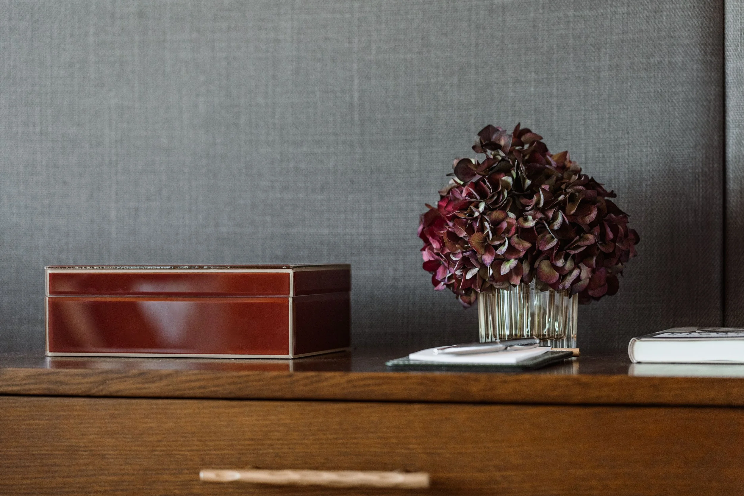 A wooden table with a red box, a glass vase with dark red hydrangea flowers, a white notebook, a pen, and a card.