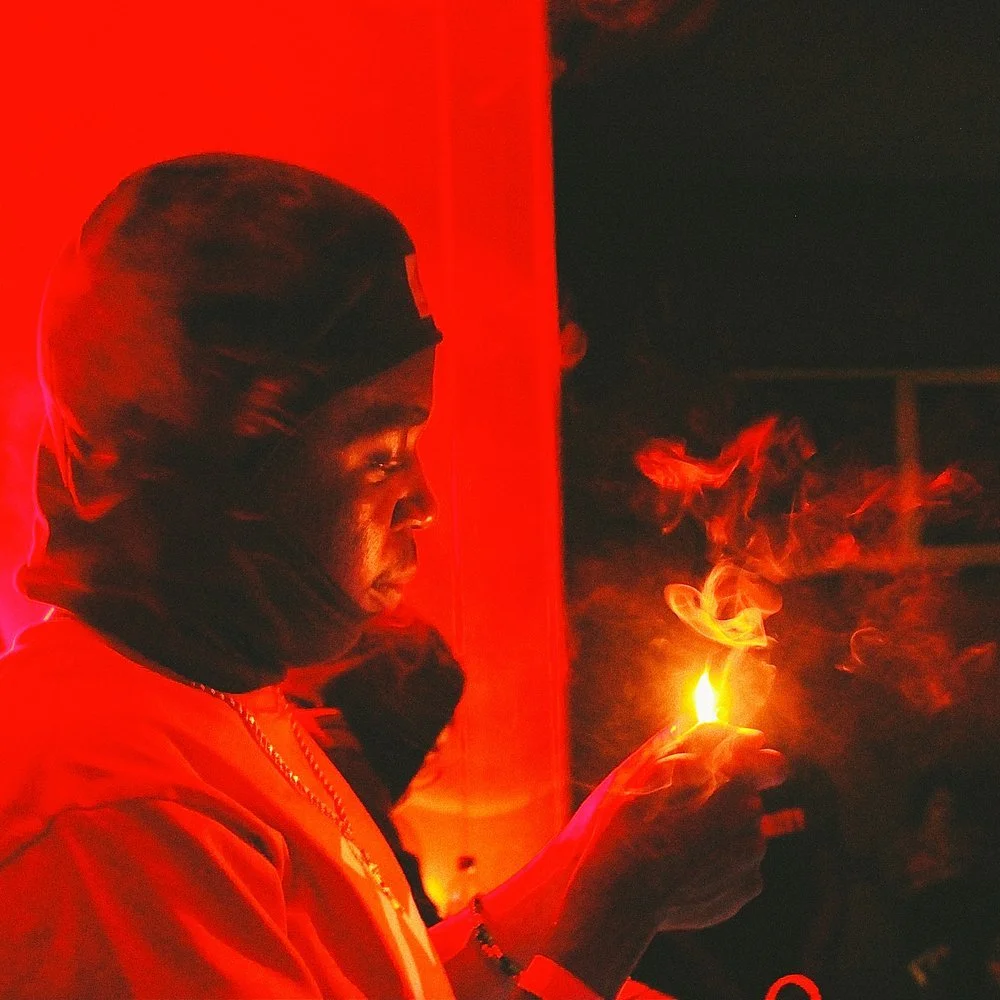 A woman holding a lit match or lighter in a dimly lit space, illuminated by a red light, with her face and hand visible and smoke rising from the flame.