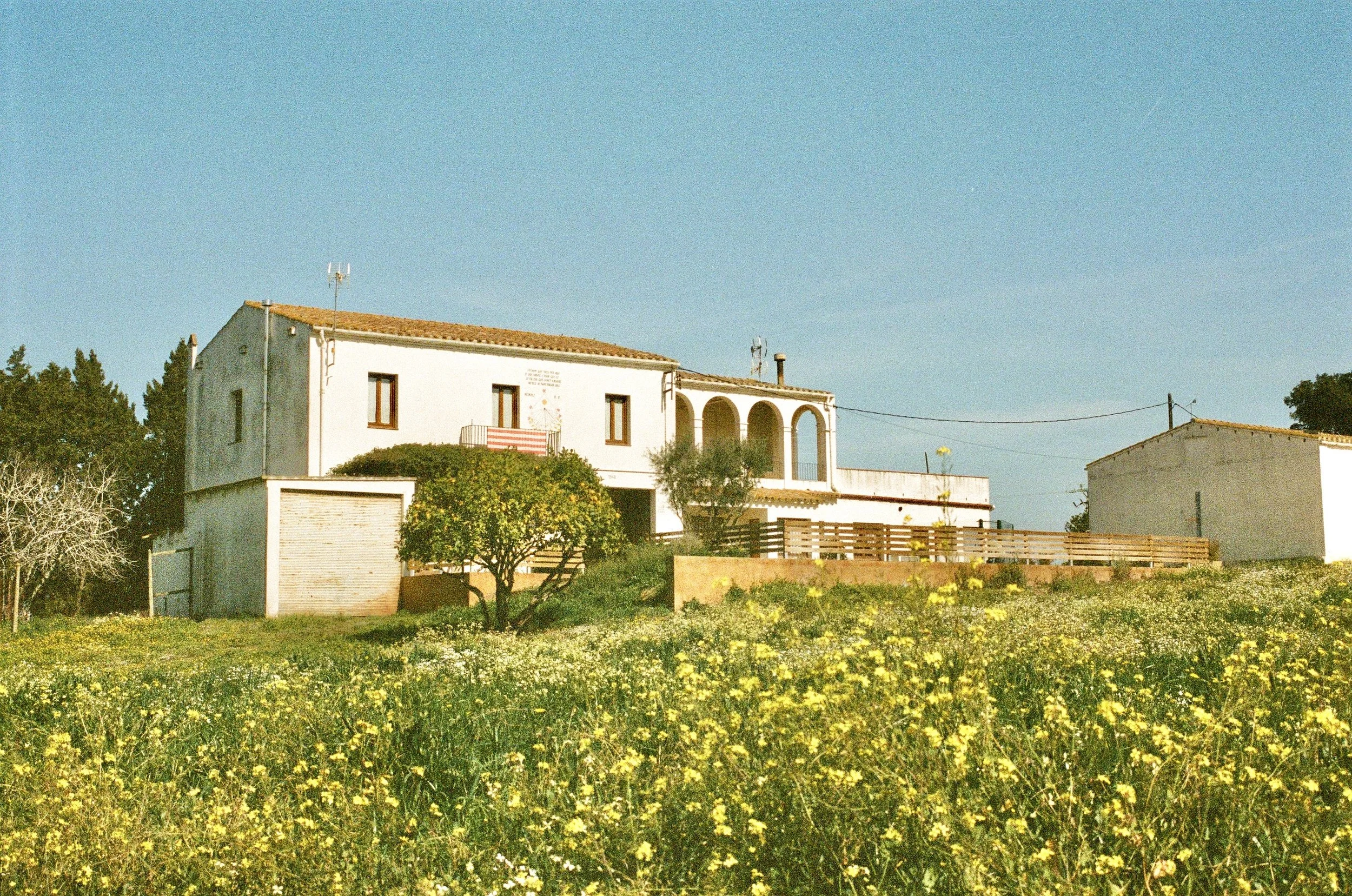 A white house with a red-tiled roof, arched balcony, and surrounding greenery under a clear blue sky.