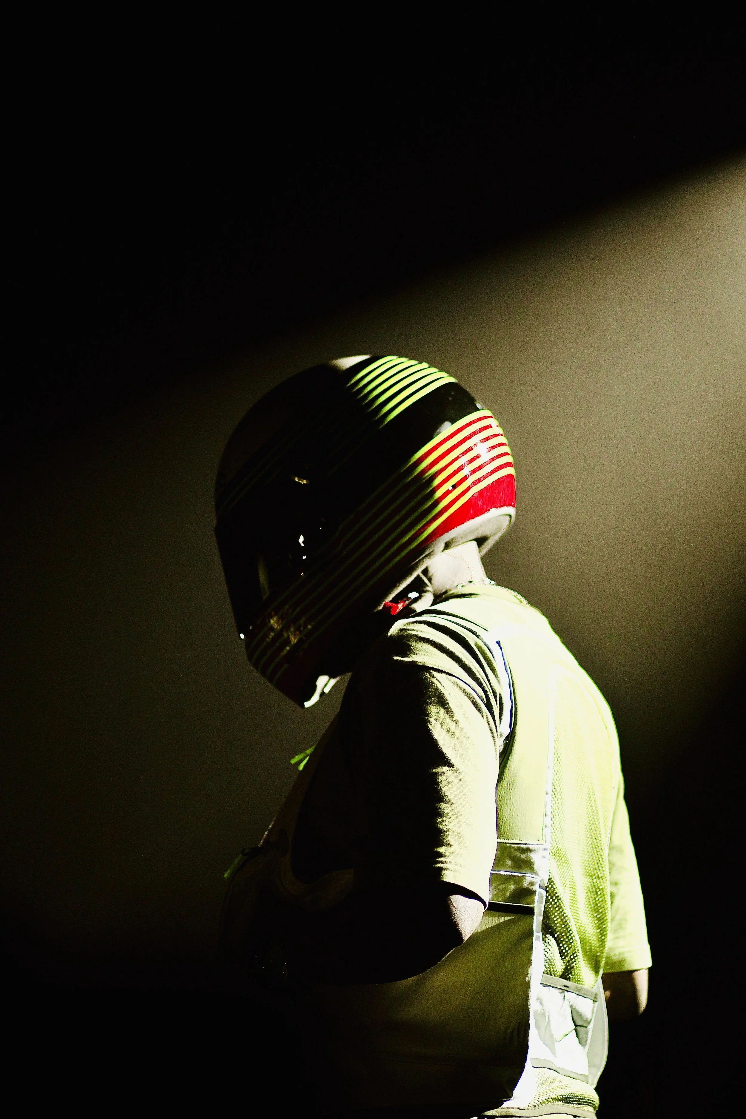 A person in a yellow safety vest and racing helmet standing in a dark environment with a spotlight illuminating part of their body and helmet.