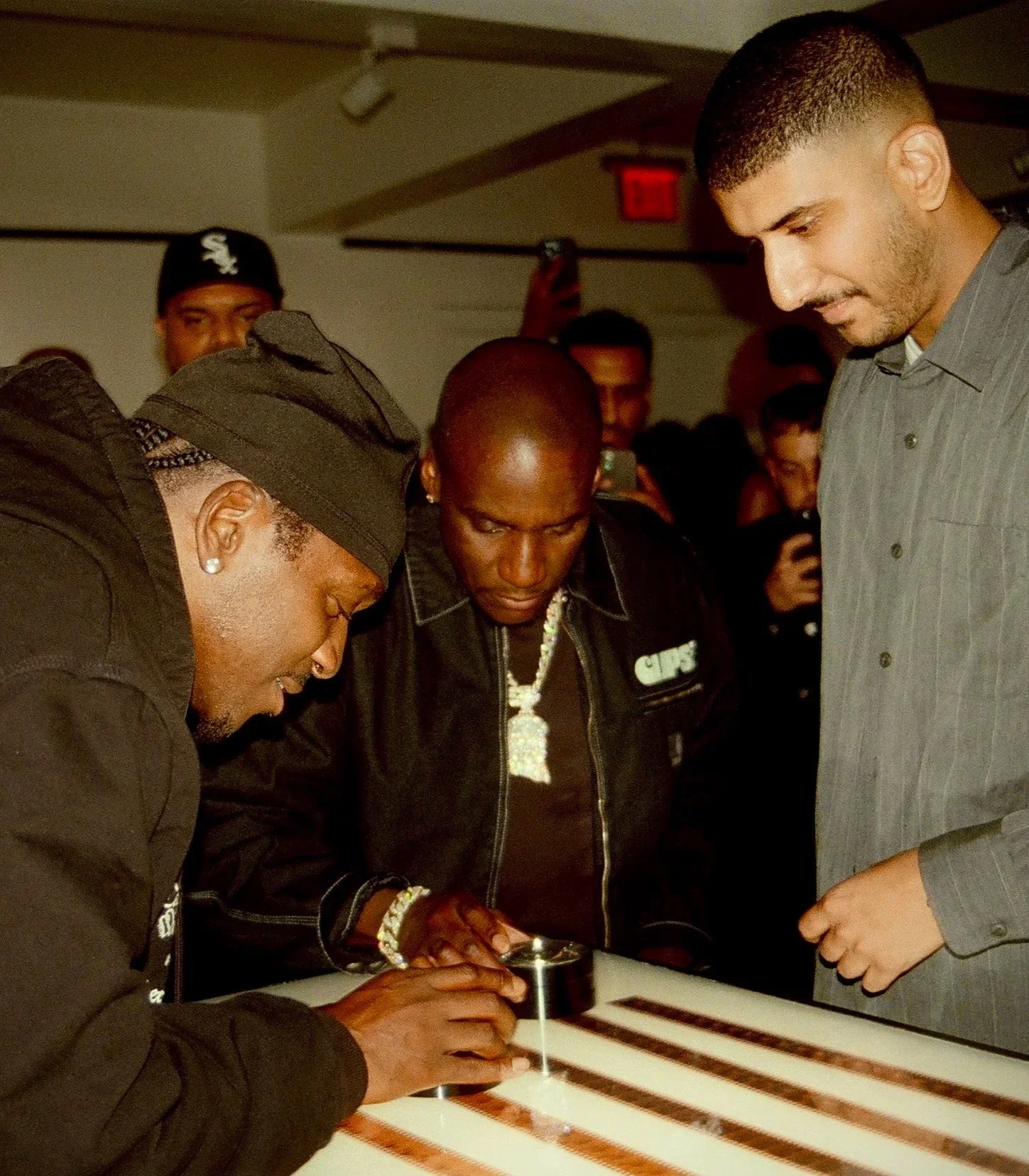 Four people are gathered around a table, one holding a magnifying glass and examining something, with others looking on. The background shows a crowd and a woman with an earring and a hat.