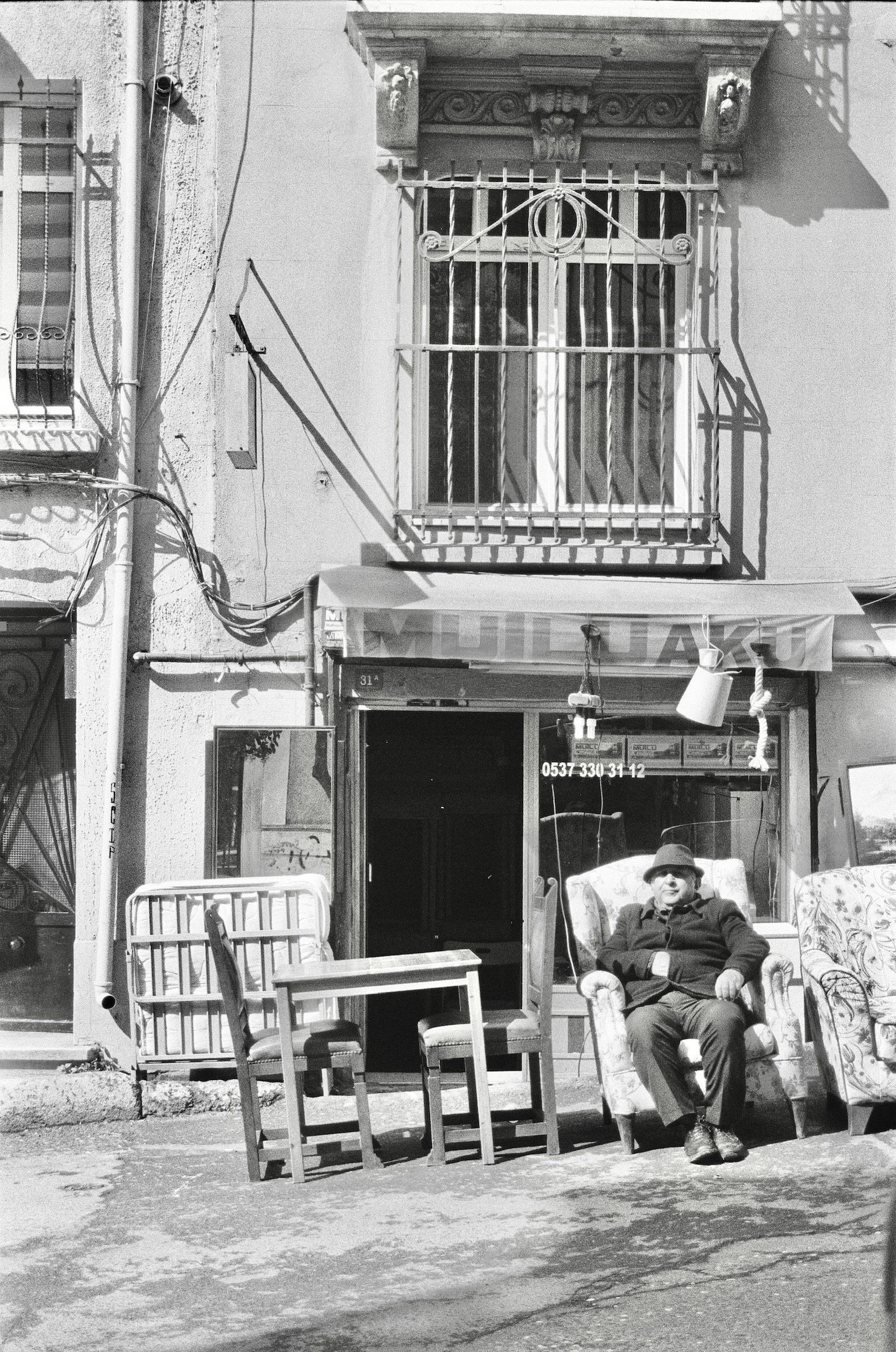 A man sitting in an armchair outdoors in front of a building with wrought iron window bars. There are various chairs and tables around, and a building with a window and an awning above the open door.