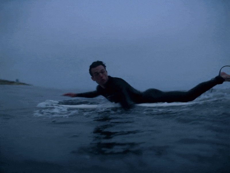 A person lying on a surfboard in the water during dusk or dawn, holding onto the board with hands and legs extended behind, with a shoreline visible in the background.