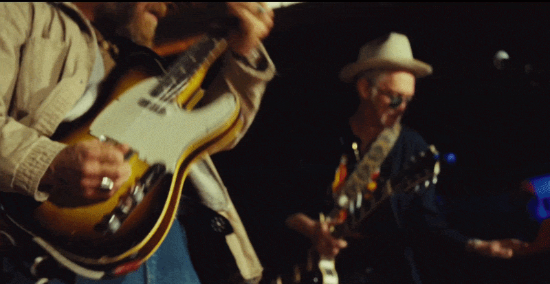 Two musicians playing guitars on stage, one with a white fedora hat, the other with sunglasses.