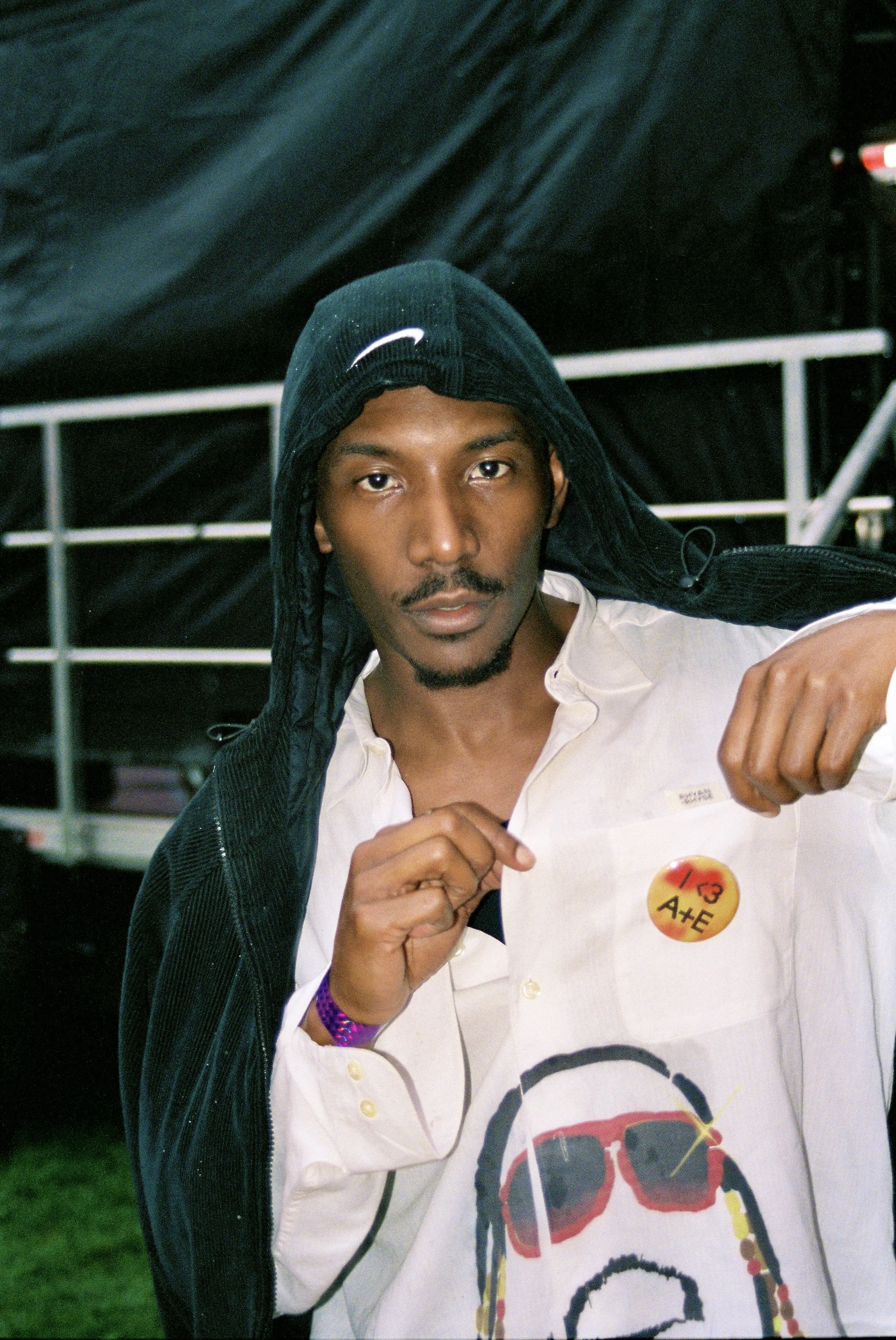 Young man wearing a black hoodie with a Nike logo, a white shirt with a design, and a purple wristband, making a fist gesture in front of a black backdrop.