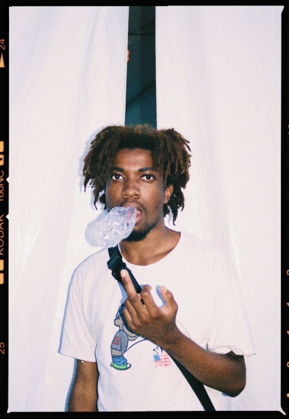 Young man with dreadlocks drinking from a plastic bottle, wearing a white t-shirt with a cartoon graphic, against a white backdrop.