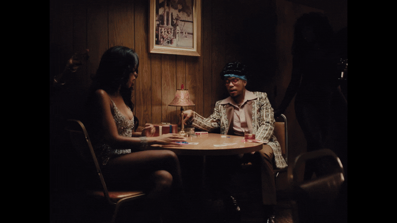 Two women are playing cards at a table in a dimly lit room with wood-paneled walls. One woman has long dark hair and is wearing a sparkly dress, while the other has short hair, glasses, and is dressed in vintage style clothing. A small table lamp with a pink shade provides light, and a framed picture hangs on the wall.