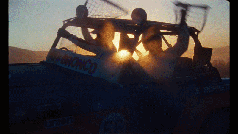 Two people in a car during sunset, with a roll cage and racing gear, celebrating.
