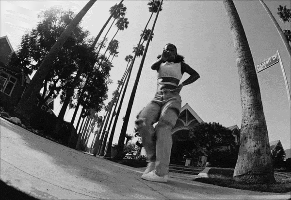 A woman is dancing outdoors on a sidewalk lined with tall palm trees, wearing sunglasses and casual clothing.