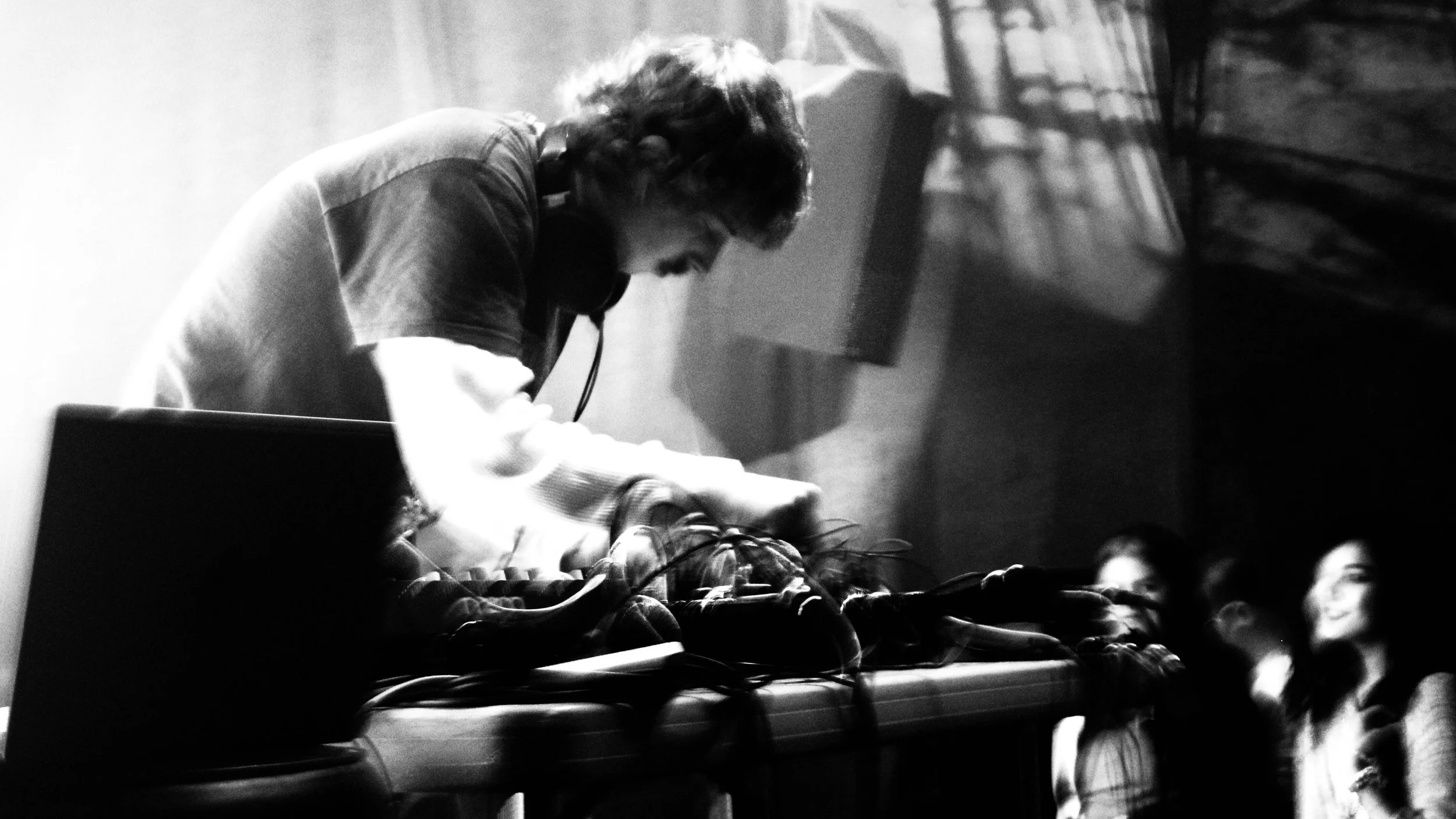 A black and white photograph of a person with curly hair wearing headphones, working with audio equipment, with a group of people watching nearby.