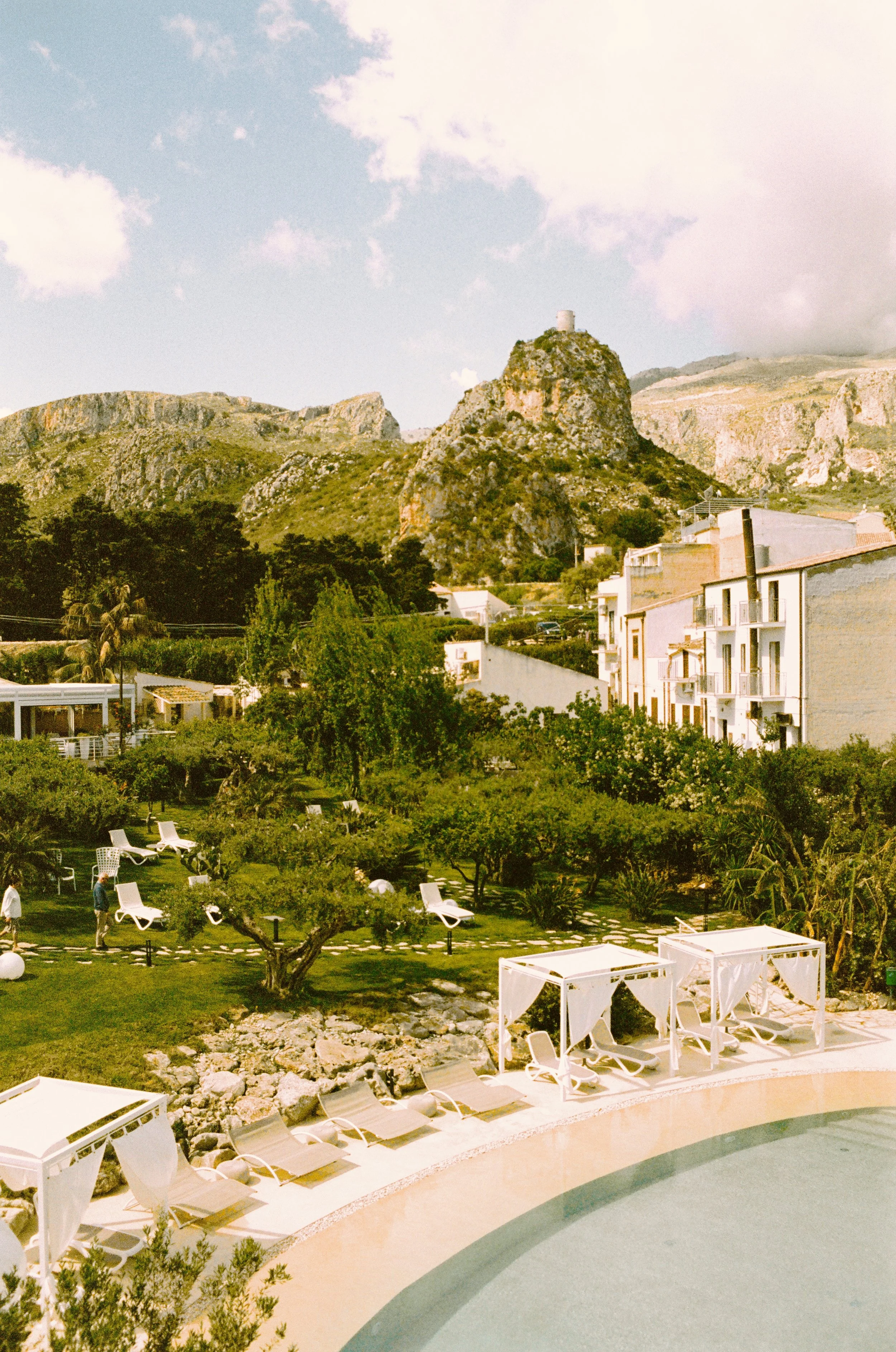 View of a hillside with rocky cliffs, greenery, and a small white building on a hilltop. In the foreground, there is a pool area with white lounge chairs and cabanas, some people walking and relaxing.