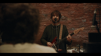 A musician with brown hair singing into a microphone and playing an electric guitar, in front of an audience with a brick wall background.