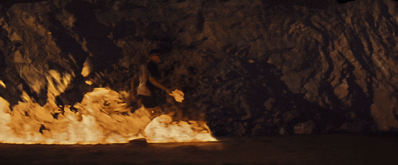A person sitting on the beach near the water at night with a fire and rocky cliffs in the background.