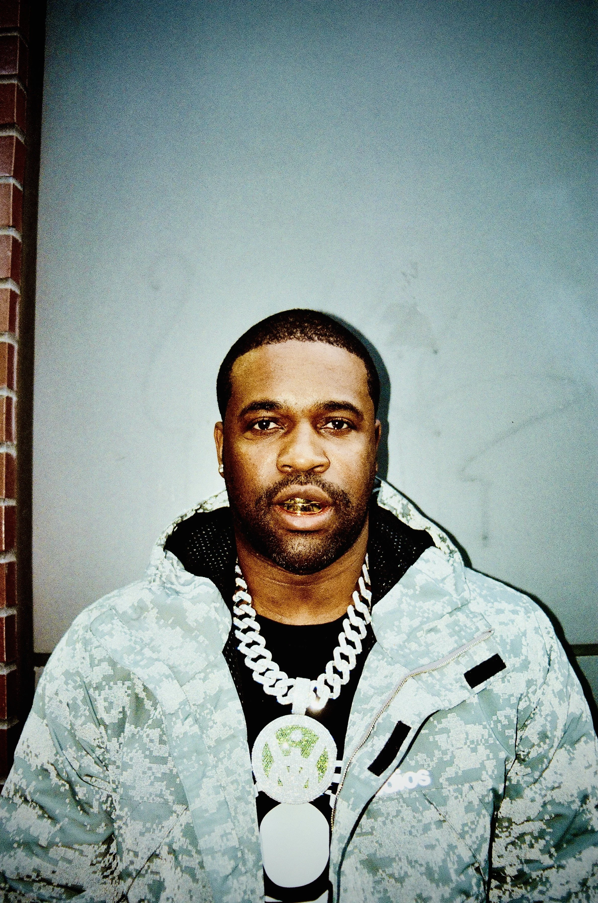 A man with dark skin, short hair, and a mustache, wearing a light-colored camouflage jacket, a black shirt with a white graphic, a large white chain necklace, and a gold grill on his teeth, standing in front of a plain gray wall.
