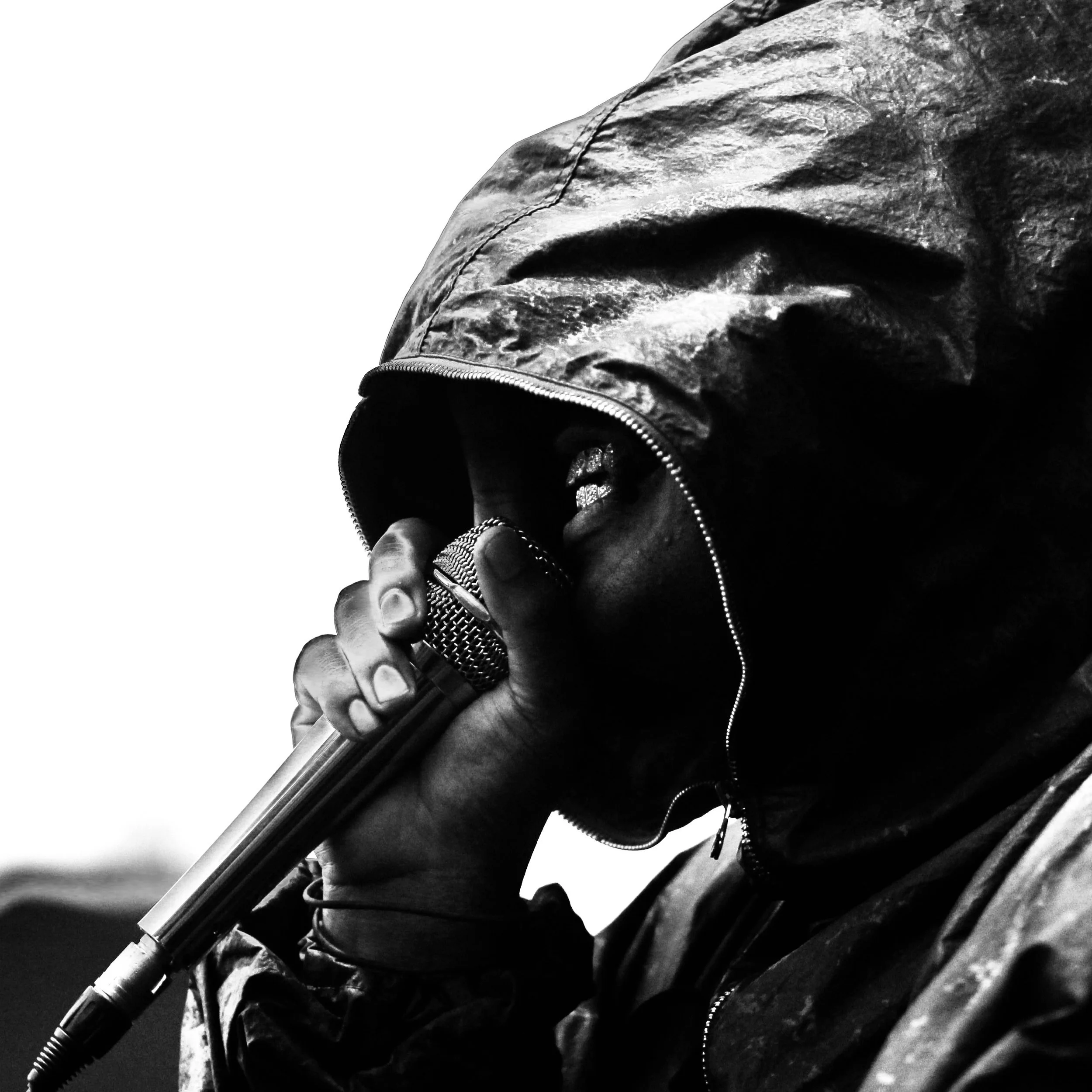 Close-up of a person's face partially obscured by a hoodie, holding a microphone close to their mouth, in black and white.