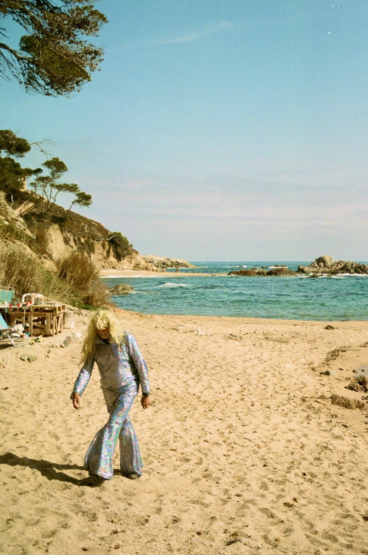 A woman walking on a sandy beach with rocky cliffs and trees in the background, under a mostly clear blue sky.