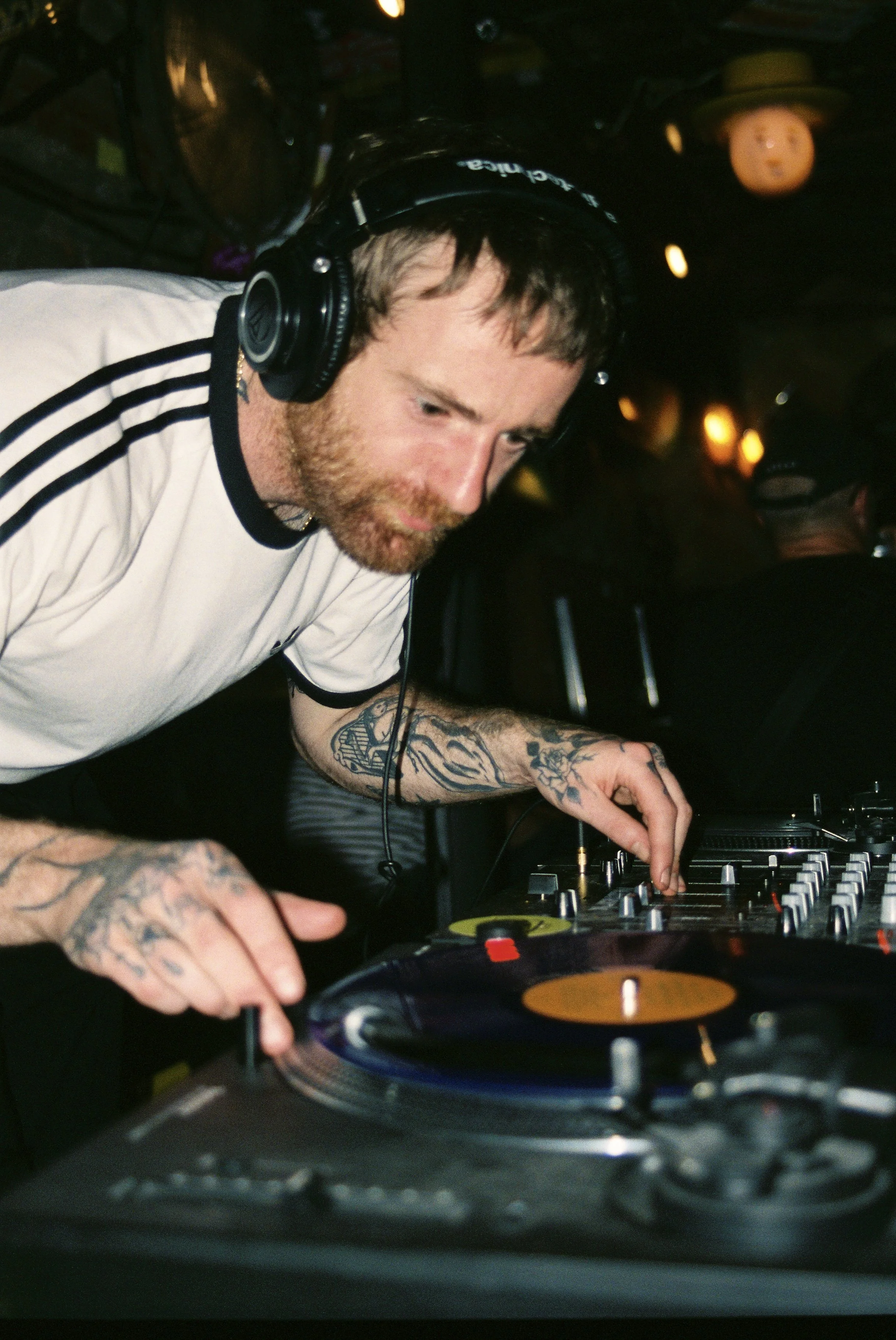 A male DJ with tattoos on his arms wearing a white t-shirt and black headphones, mixing music on DJ equipment in a dimly lit setting with floating lights and ceiling decorations.