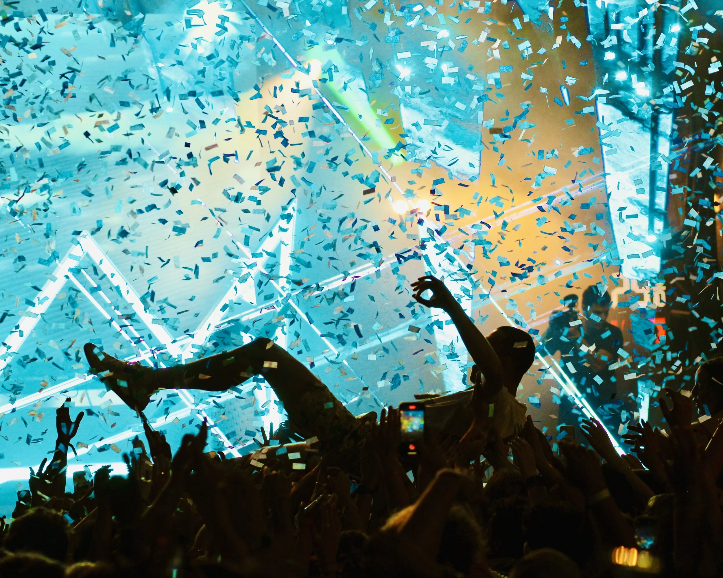 Crowd of people at a concert or celebration with blue and yellow confetti falling, illuminated by bright colorful stage lights, and some hands raised in the air.