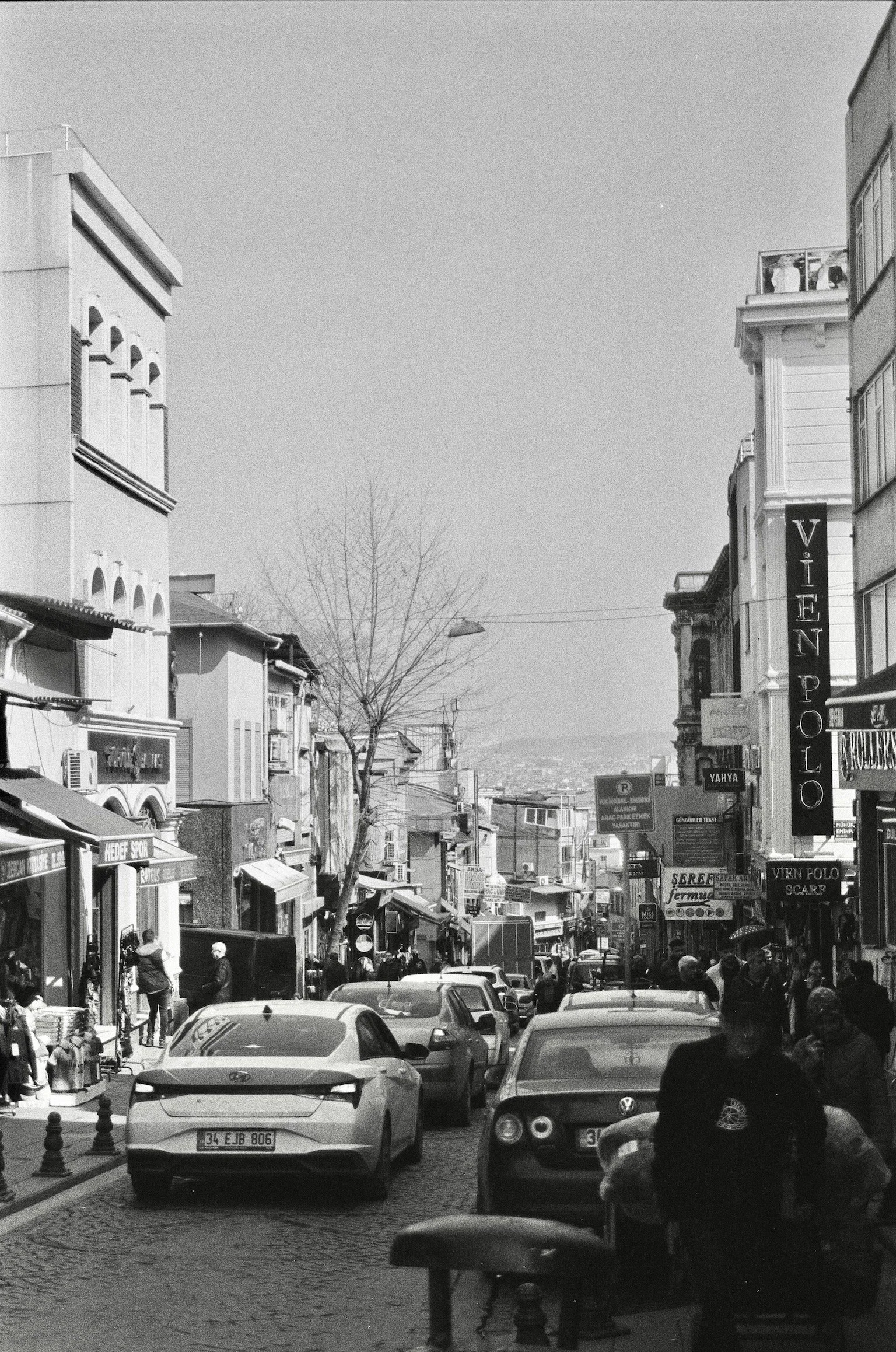 A busy street scene with parked cars, pedestrians, and storefronts, in black and white.