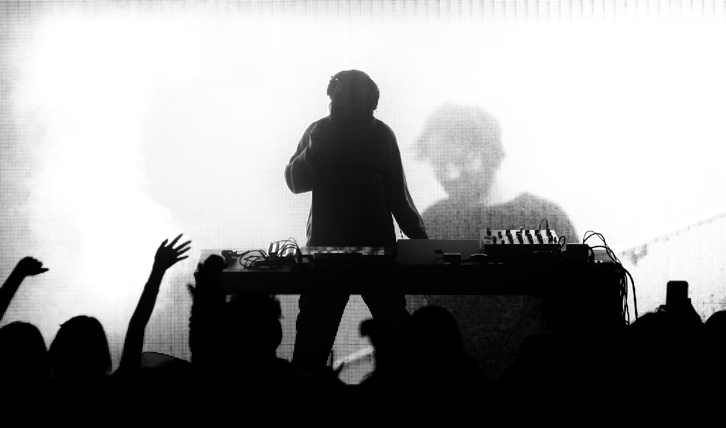 Black and white photo of a DJ performing on stage with a large screen behind him displaying his silhouette. Audience members are visible with raised hands in the foreground.