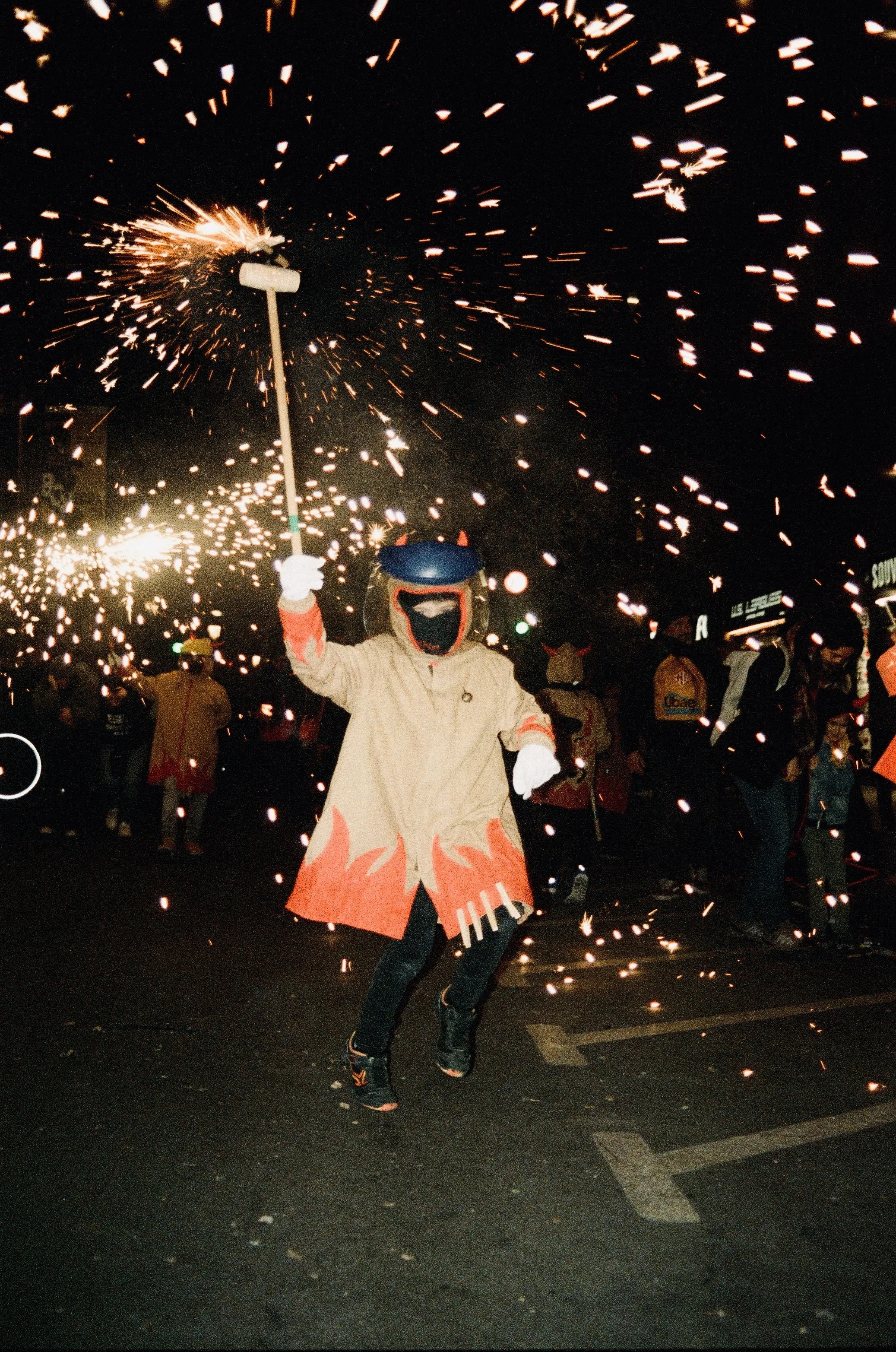 Person dressed as a devil or demon with a blue helmet, orange horns, and flame-patterned coat, holding a sparkler at night, with a crowd of people in the background.