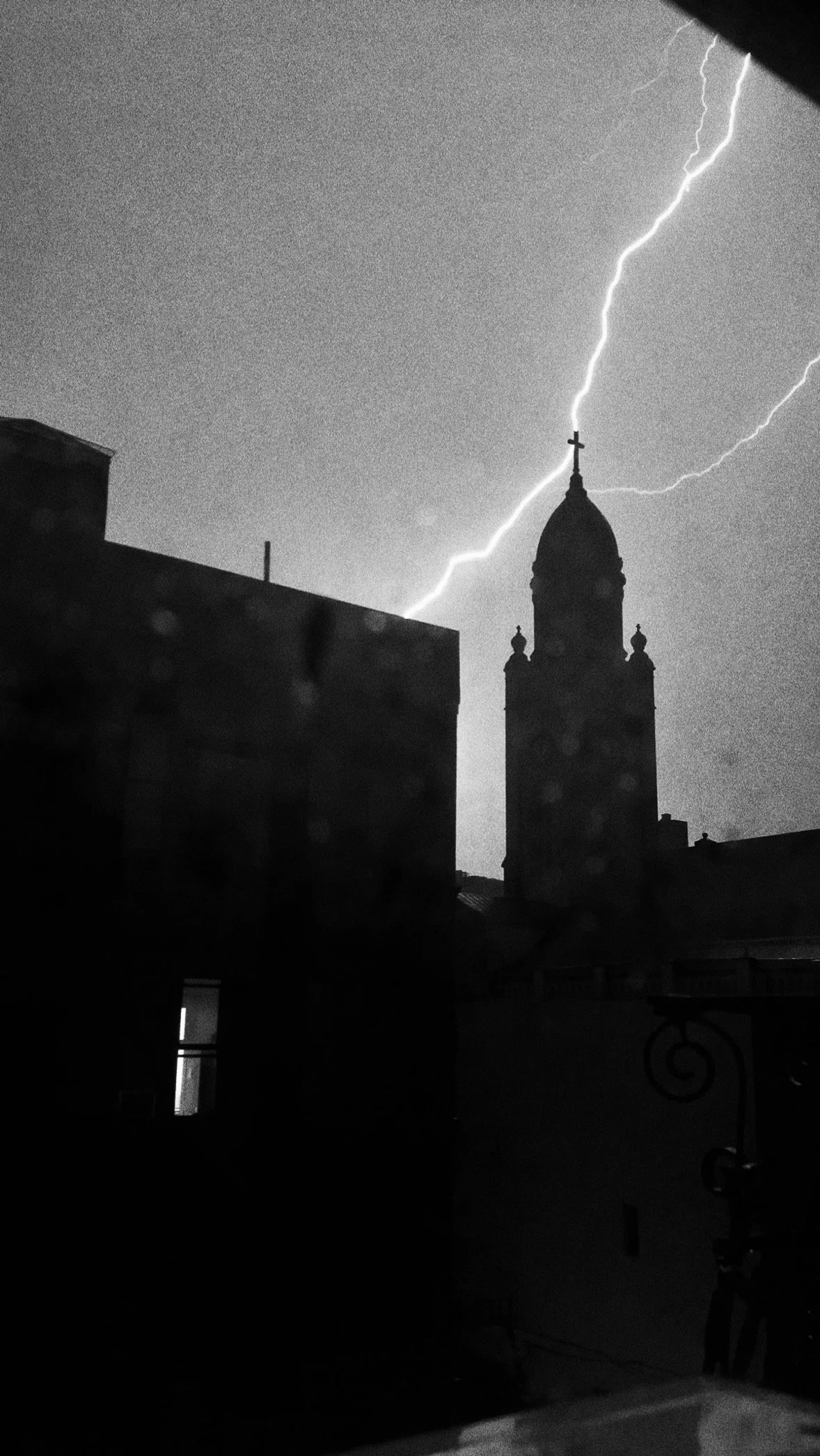Black and white photo of a lightning strike hitting the top of a building with a cross on it, overcast sky in the background, silhouette of other buildings and rooftops in the foreground.