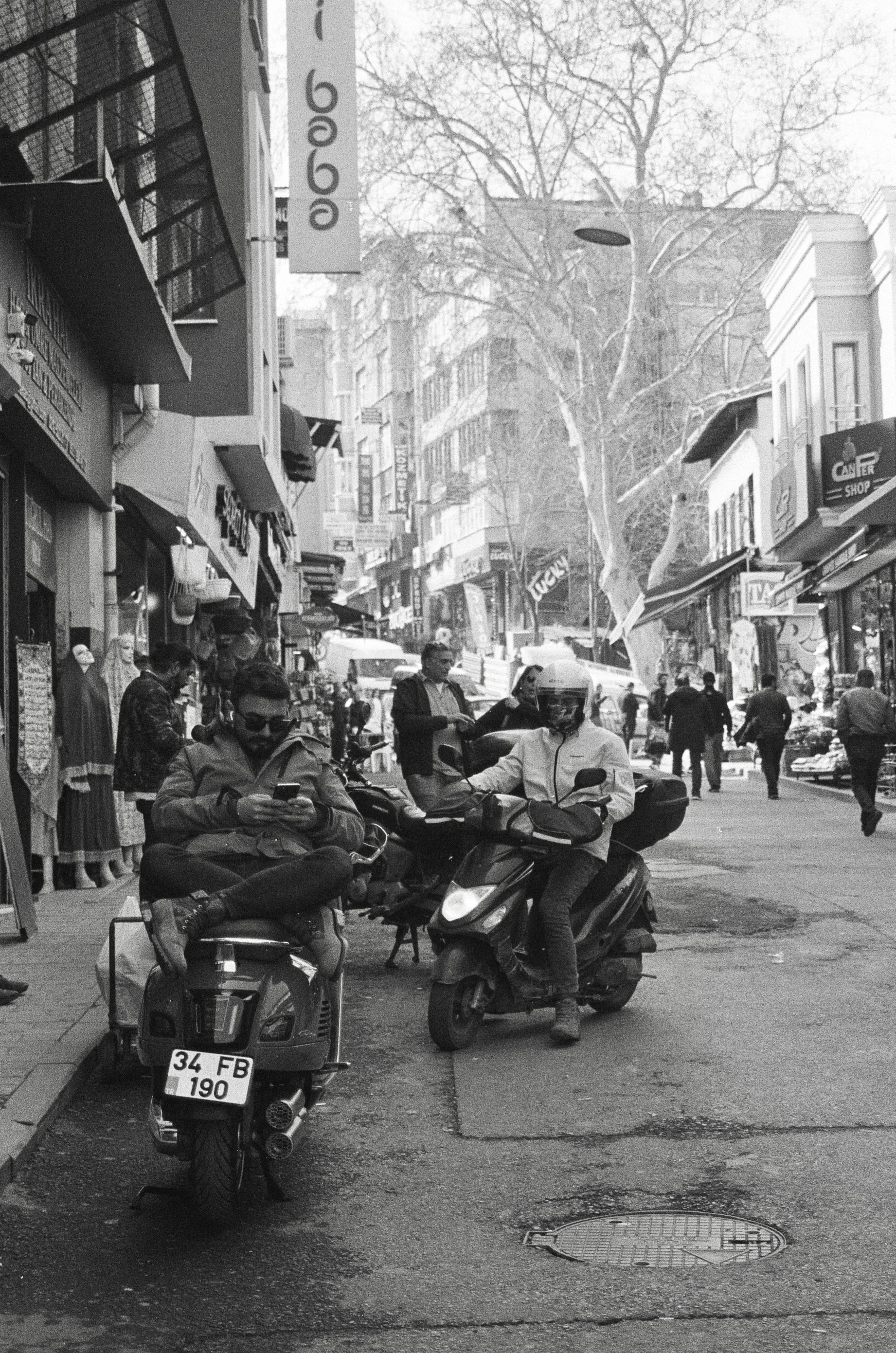 People on scooters and walking on a busy city street lined with shops and signs, with buildings and a large tree in the background.