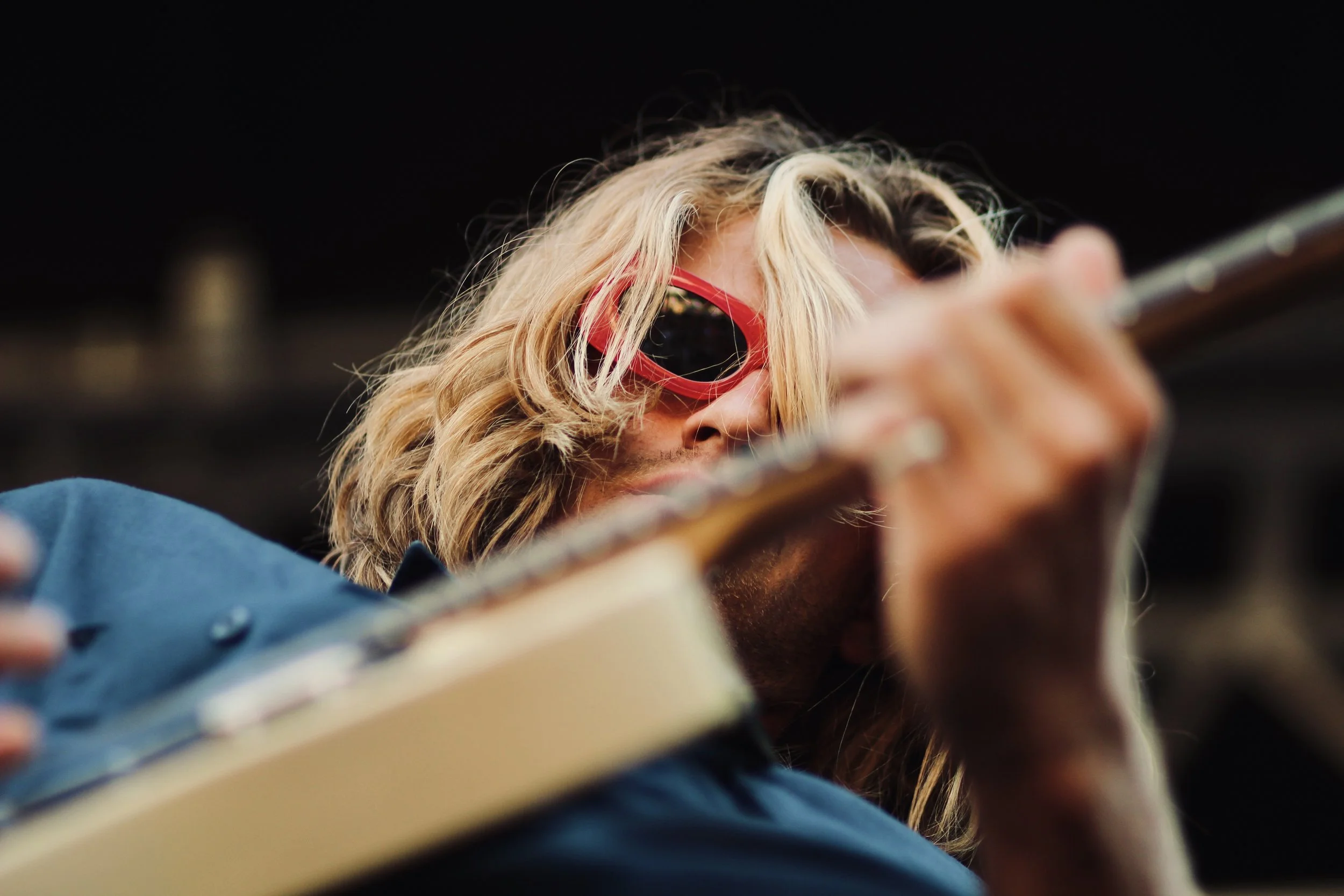 A person with blond, wavy hair wearing red sunglasses and a blue shirt, playing a guitar.